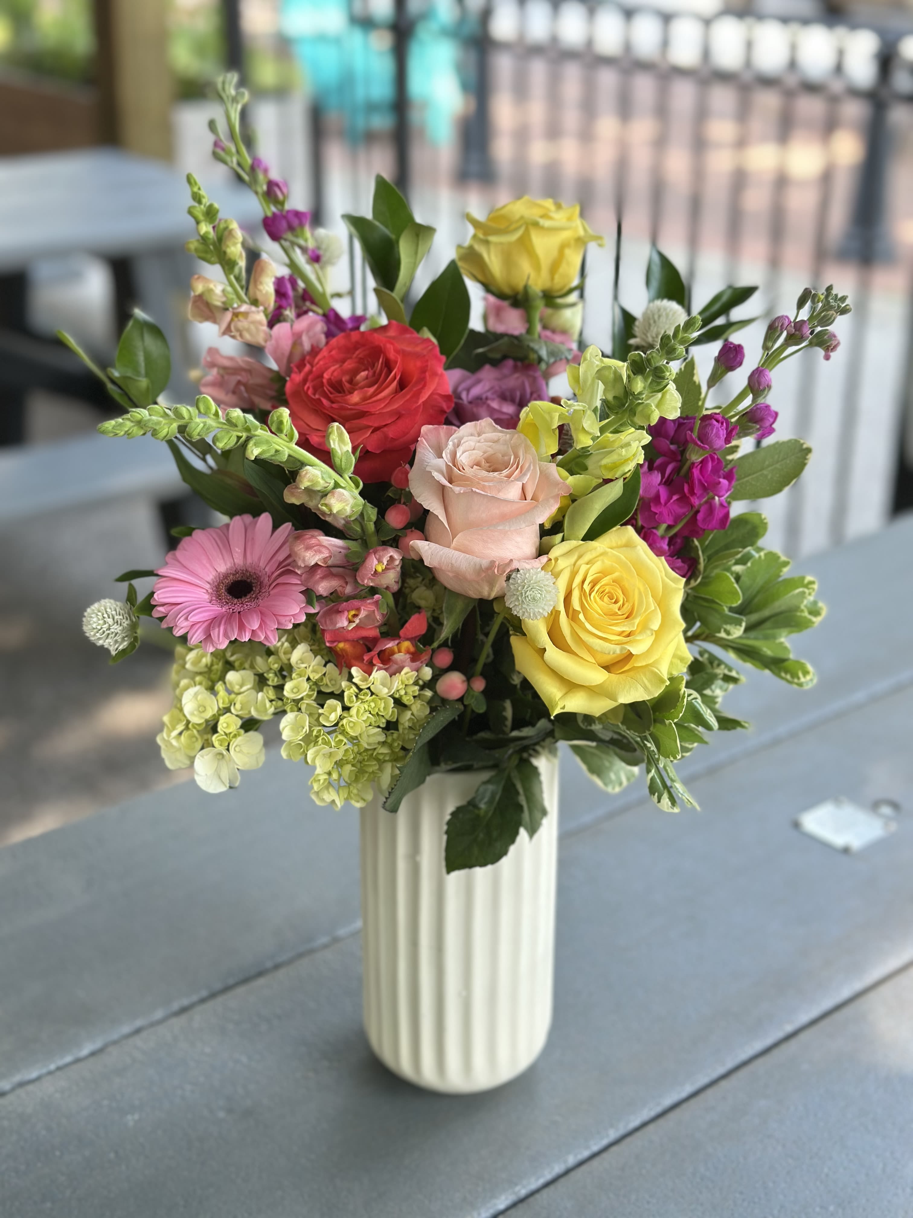 Summer Picnic - A bright, fresh-picked look that feels like sunshine in a vase - this flower arrangement is a celebratory everyday bouquet perfect for a birthday, congratulations, or a cheerful spring gift. The Summer Picnic arrangement pairs roses and snapdragons with a single pink gerbera and clustered hydrangea blooms to create a lively, garden-style display your person will notice the moment it arrives.  Colors span cheerful yellow, coral red, blush pink and vivid lavender-purple, set against glossy green foliage and small round white filler blooms for texture and movement. Flowers are gathered in a rounded, slightly airy composition and presented in a tall white ribbed ceramic vase that gives the arrangement a polished yet relaxed feel.  Send this to mark happy occasions or simply to make someone feel truly seen - it reads as upbeat and joyful, the kind of bouquet that brightens a kitchen table or office desk. The tall vase helps the snapdragons stand proud while keeping the mixed blooms comfortably arranged; minimal care beyond fresh water will keep the flowers looking fresh.