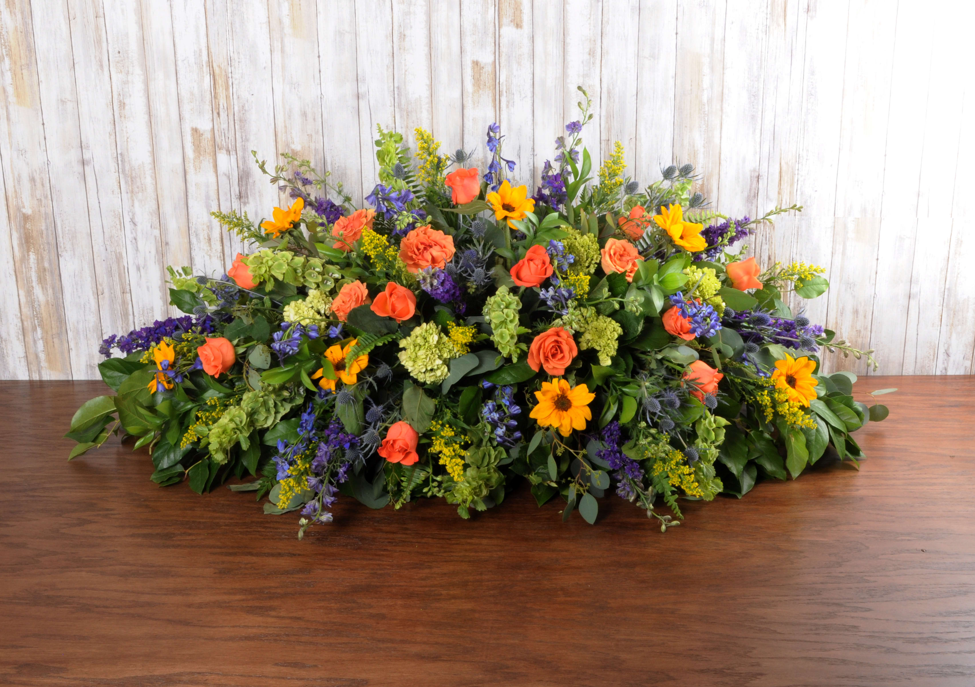 Low spray arrangement of orange roses, yellow daisies, and purple flowers in an elongated shape