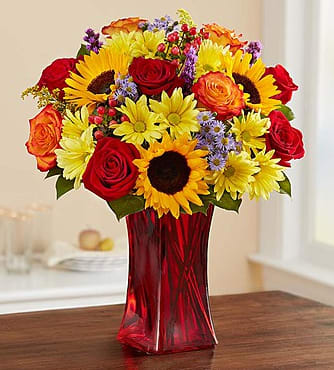 Autumn Amazement - Our brilliant new arrangement will definitely leave them delighted! Beautifully hand-designed by our expert florists, each fall-toned bloom is highlighted by a sleek ruby red gathering vase. Whatever sentiment you’re trying to capture, this warm and wonderful surprise is one they won’t soon forget. Details: All-around arrangement of red roses, carnations and hypericum, circus or bi-color orange roses, sunflowers, yellow daisy poms, purple liatris and monte casino; accented with solidago and salal. 