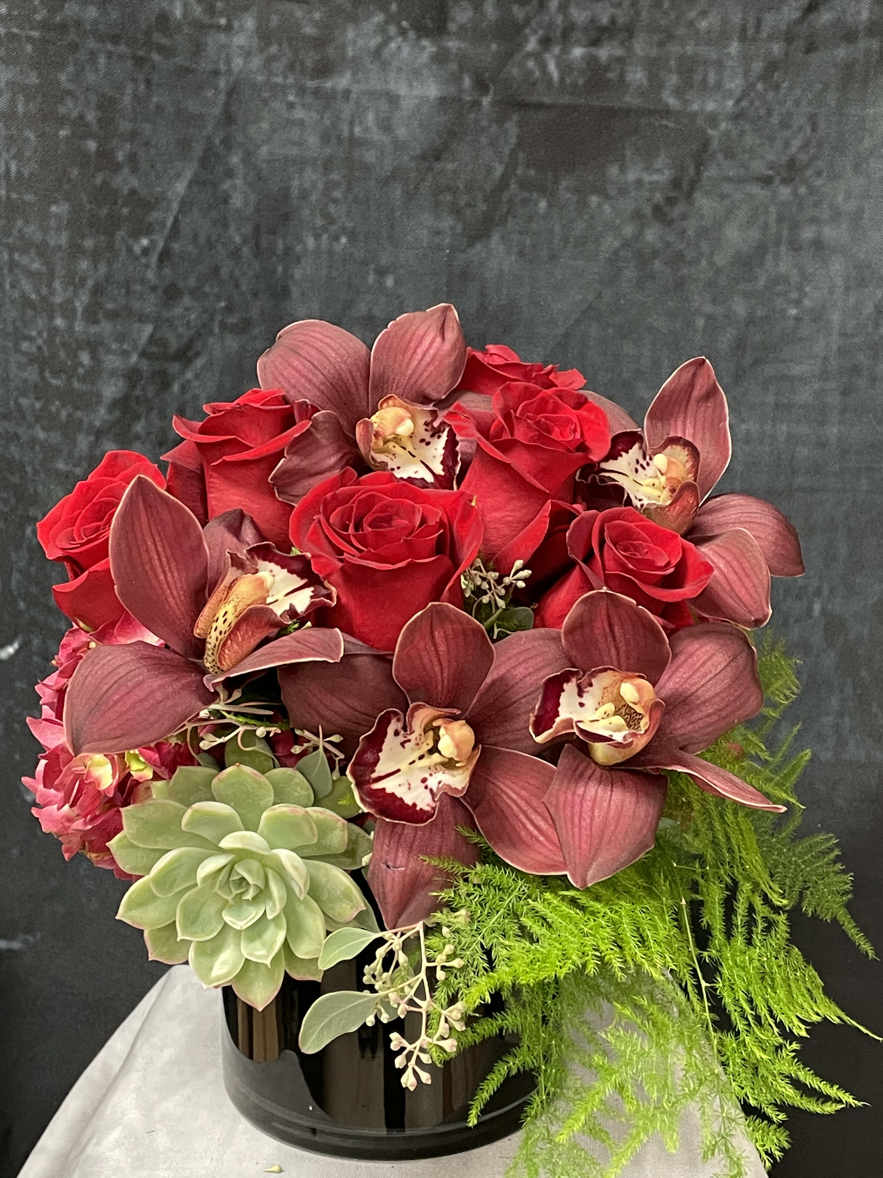  La Vie En Rouge - Natural red hydrangeas, red roses, burgundy cymbidiums, succulent and greenery in a black short cylinder vase. 