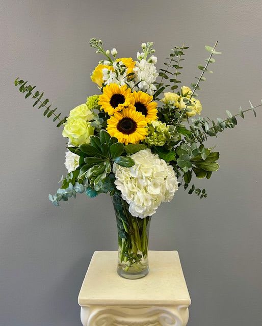 Sunflower Elegance - This striking design is suitable for all occasions. A tall glass vase is filled with eucalyptus and gorgeous yellow, white, and pale green flowers featuring groupings of sunflowers.