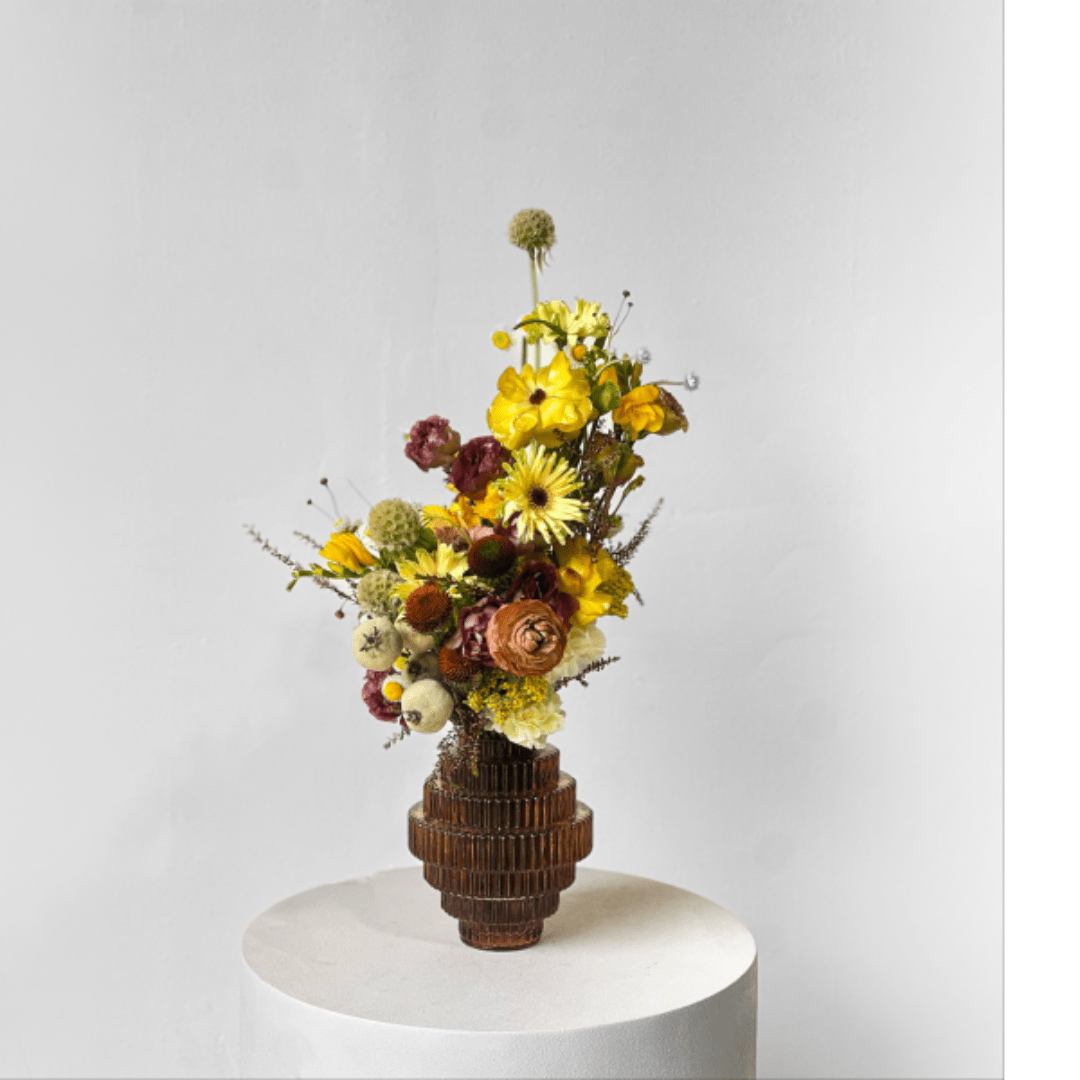 Tall mixed flower arrangement in a ribbed brown vase on a white pedestal