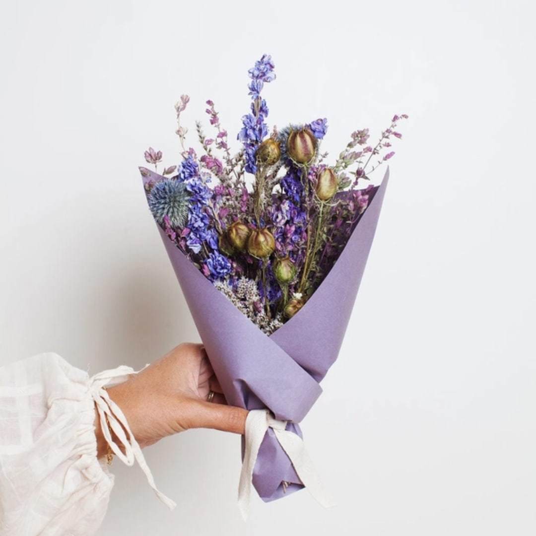 Handheld bouquet of dried purple and blue flowers wrapped in lavender paper