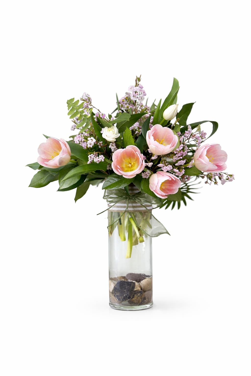 Tulip Garden - Soft pink tulips with pale yellow centers open like a secret in this springtime flower arrangement, a gentle birthday bouquet or Mother's Day gift that will make the person you're thinking of feel truly seen. The Tulip Garden pairs tulips with tiny lavender filler blooms and a few small white rose buds for a soft, romantic palette of pink, lavender, white and fresh green-perfect for sending warm wishes, celebrating a milestone, or brightening a bedside table.  Gathered in a tall clear glass cylinder vase, the stems sit above smooth decorative stones visible through the water and are finished with a sheer ribbon tied around the neck for an airy, elegant touch. The relaxed, natural shape and seasonal color mix read as both modern and effortless; the arrangement stands about the height of the vase and fills a small-to-medium space without overwhelming it. Shipped from our Lewiston, NY studio, each Tulip Garden is hand-crafted to reflect the same light, fresh style-substitutions may be used to maintain quality and color harmony-so she'll receive a bouquet that looks as thoughtful as the sentiment behind it.