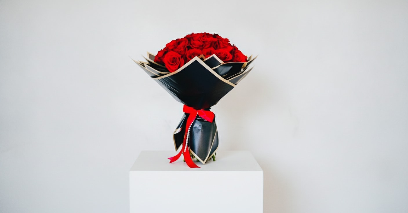 Camille Rouge ( 50, Premium Red Roses Bouquet) - A dense dome of premium red roses creates an immediate, passionate impression-perfect as an anniversary bouquet or for any love and romance moment. The vivid crimson blooms are packed into a compact, luxurious cluster with a glossy, velvety finish that reads as both opulent and intimate; wrapped in structured black paper with a light-gold border, the dramatic contrast frames the roses like a jewel. A bright red satin ribbon and a strand of metallic beads finish the handle area, giving the gift a polished, celebratory feel ideal for anniversaries, Valentine's Day, or milestone romantic gestures.  This bouquet is a statement of devotion: the tight, rounded form and rich red hues convey deep affection while the black-and-gold wrap adds a modern, elegant edge. Sizes are intentionally compact and luxurious, designed to sit beautifully in the hands or a vase; for best enjoyment, keep stems in fresh water, away from direct heat, and trim stems slightly every few days to preserve their lush, long-lasting presence.