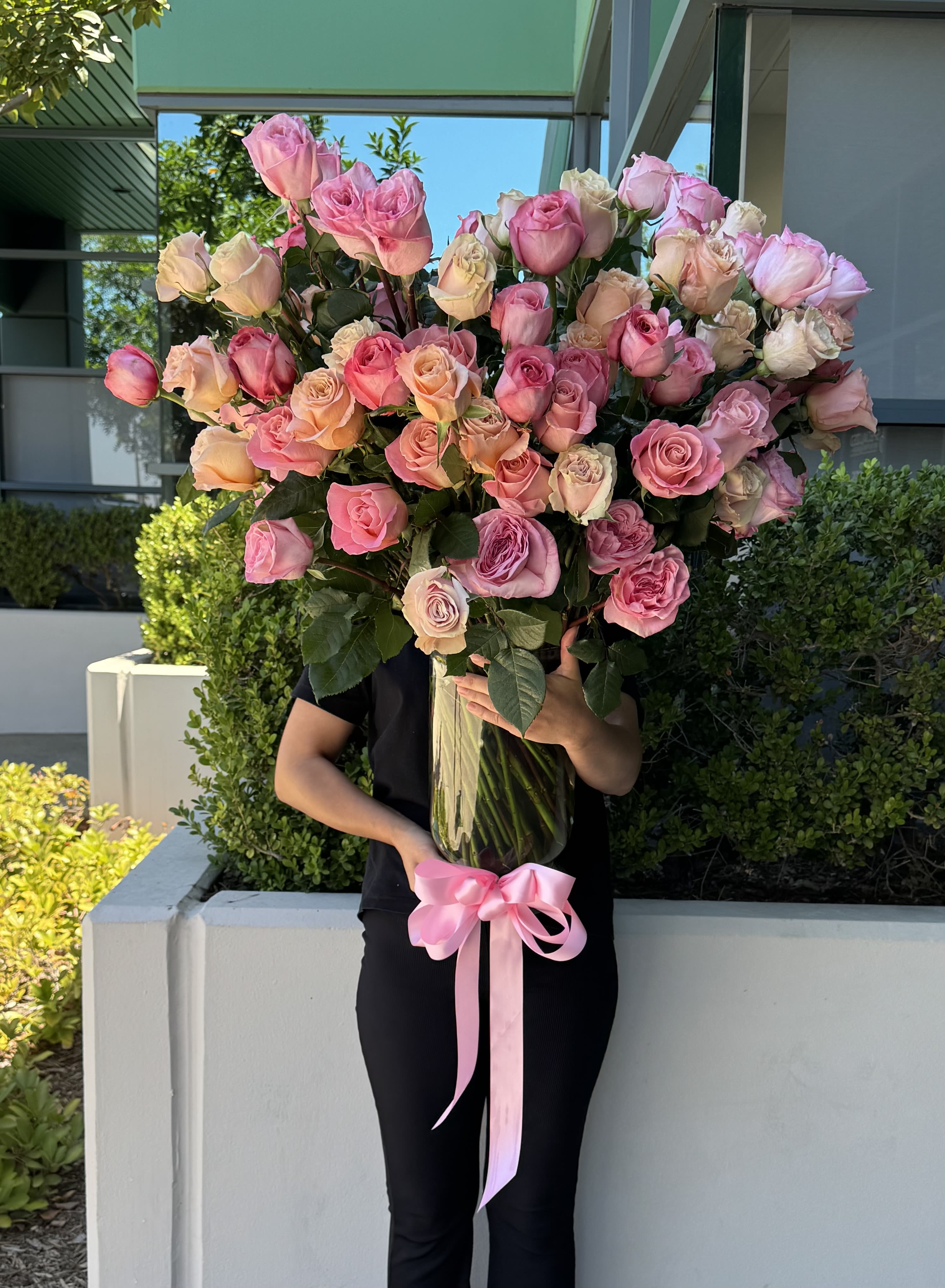 Large tall vase of pink and peach roses tied with a pink ribbon, held by a person outdoors