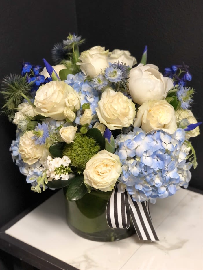 Cream roses and blue hydrangeas arranged in a glass vase with a striped ribbon.