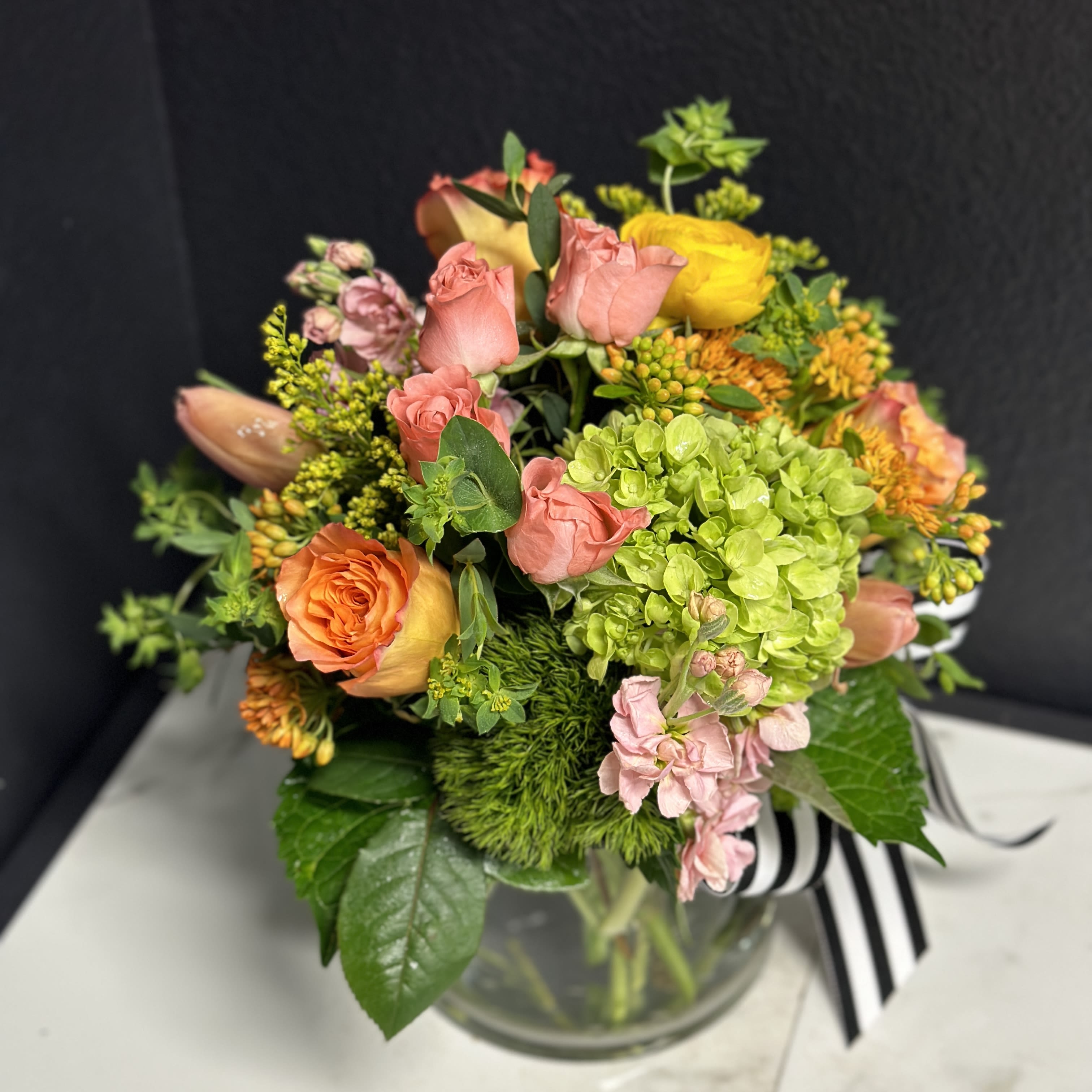 Mixed bouquet of pink, orange, yellow, and green flowers in a glass vase