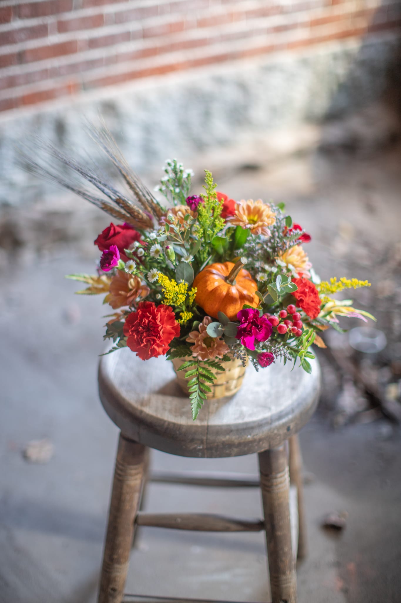 Petite Pumpkin - A mini pumpkin gourd pick is the focal point of the arrangement. It is nested around fall jewel tone blooms. This bamboo basket is complete with seasonal bearded wheat. Original design by Katie. 