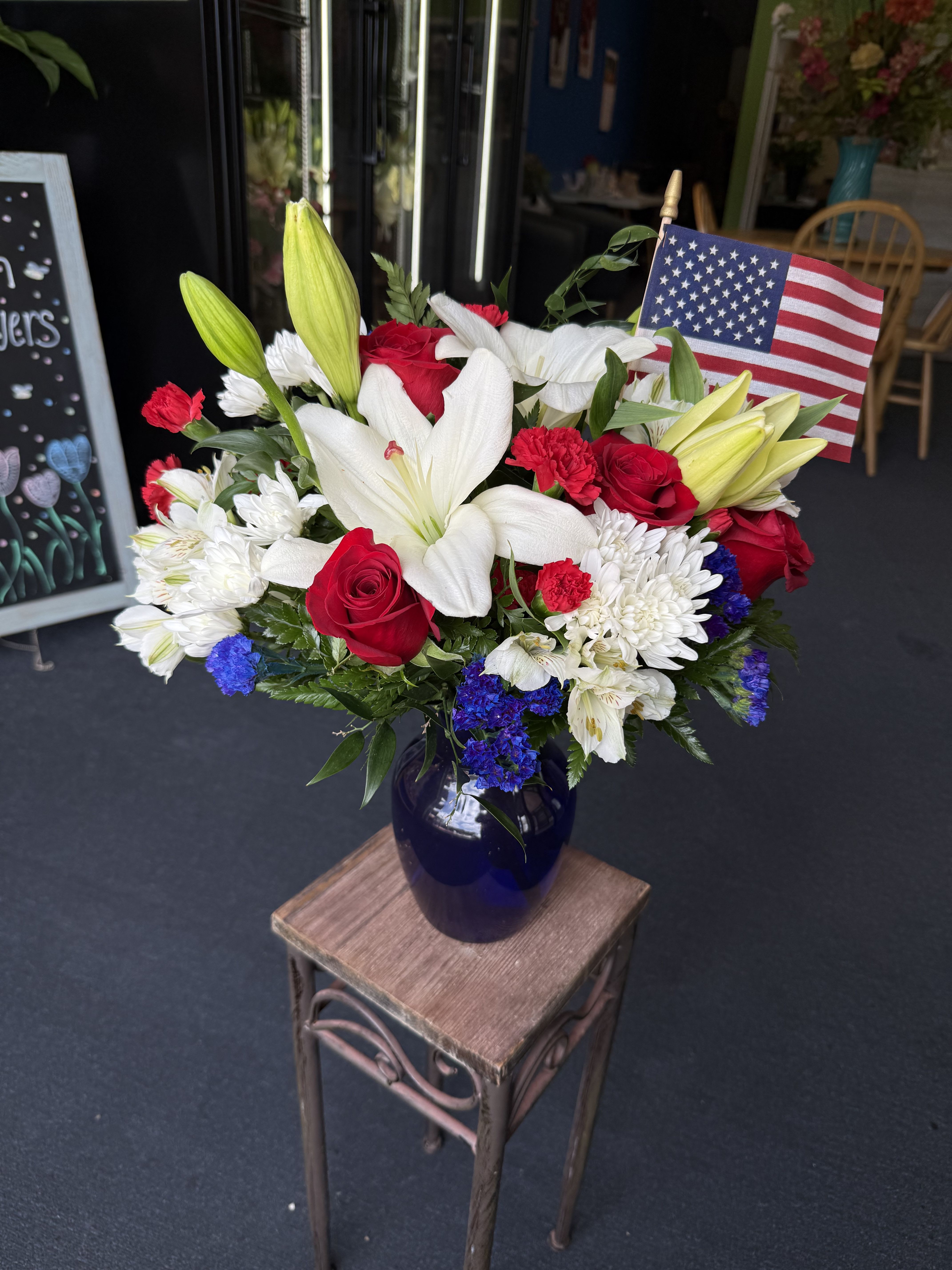 In honor  - Patriotic red, white and blue arrangement of white lilies, red roses, carnations, chrysanthemums and blue fillers in a deep blue vase with flag accent-bold, full, and ceremonially fresh.