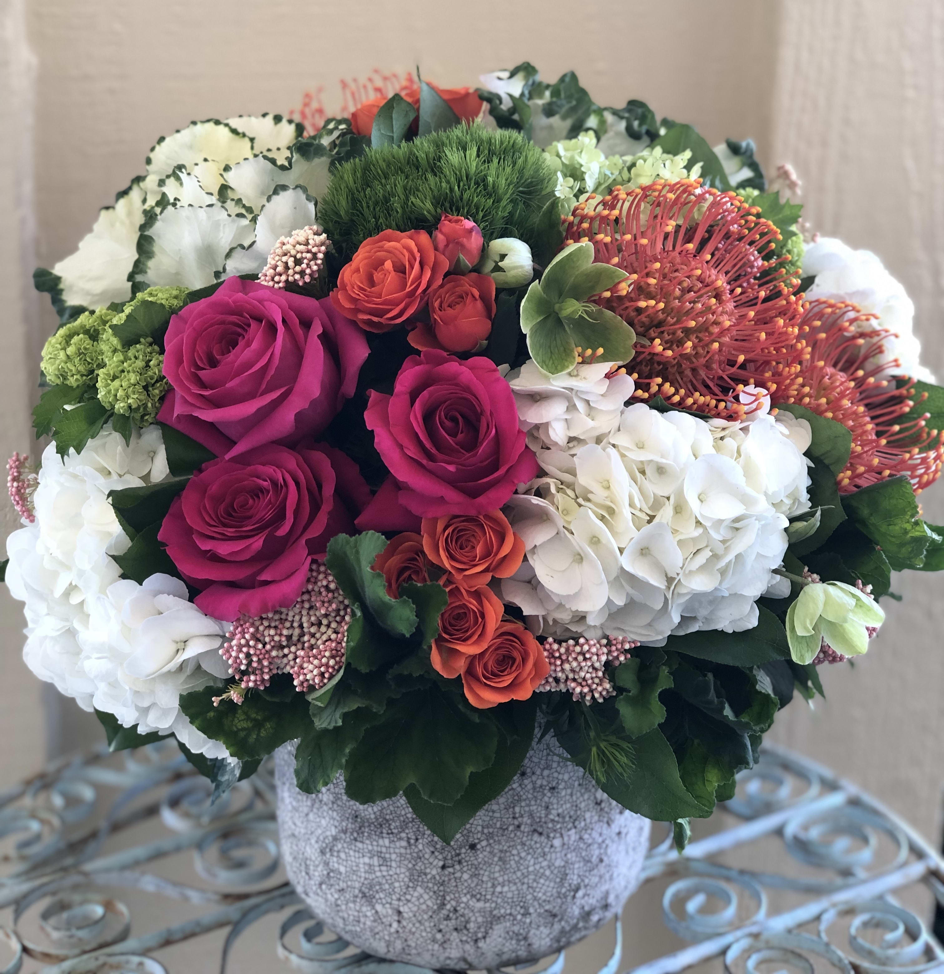 Low round arrangement of pink roses, orange spray roses, protea, and white hydrangeas in a gray pot