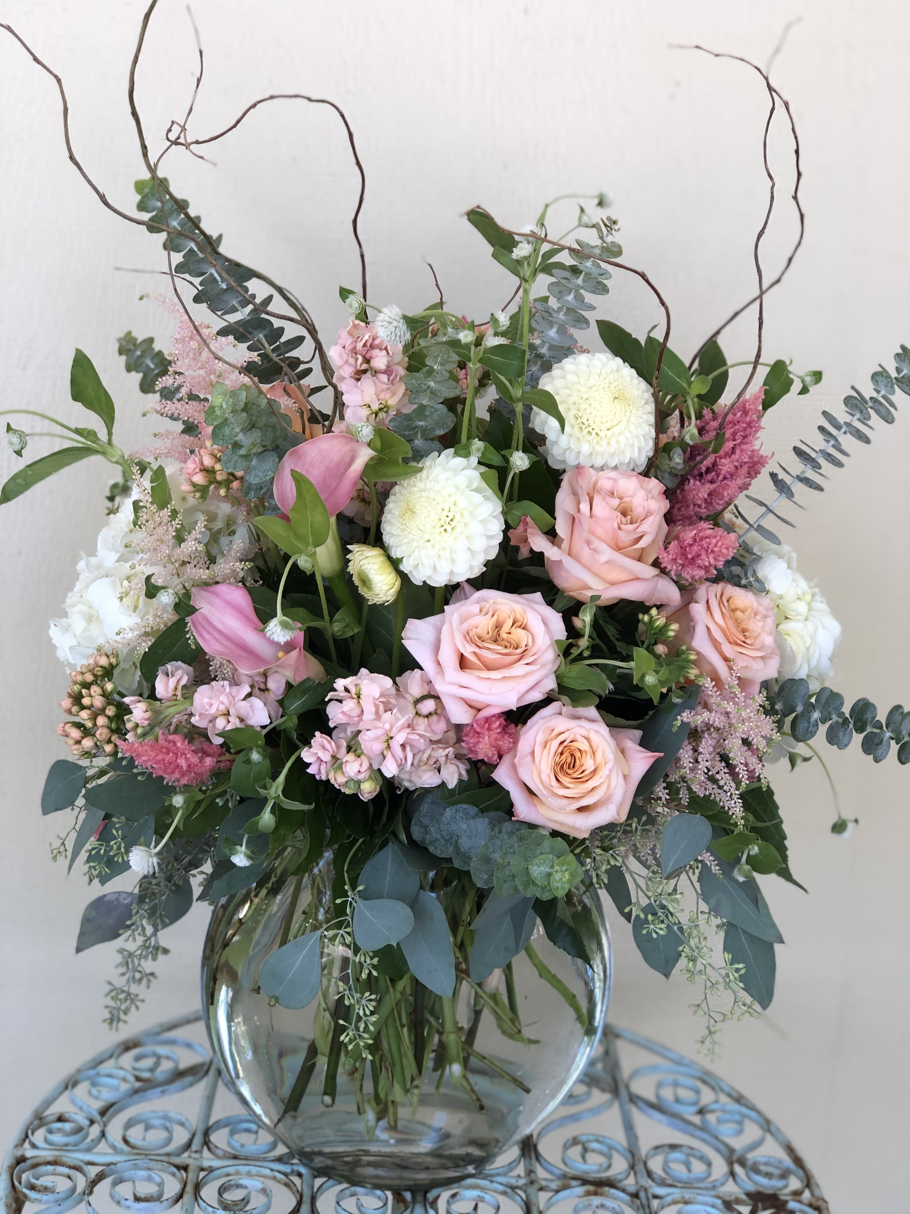 Tall pastel arrangement of pink roses, white mums, and calla lilies in a round glass vase