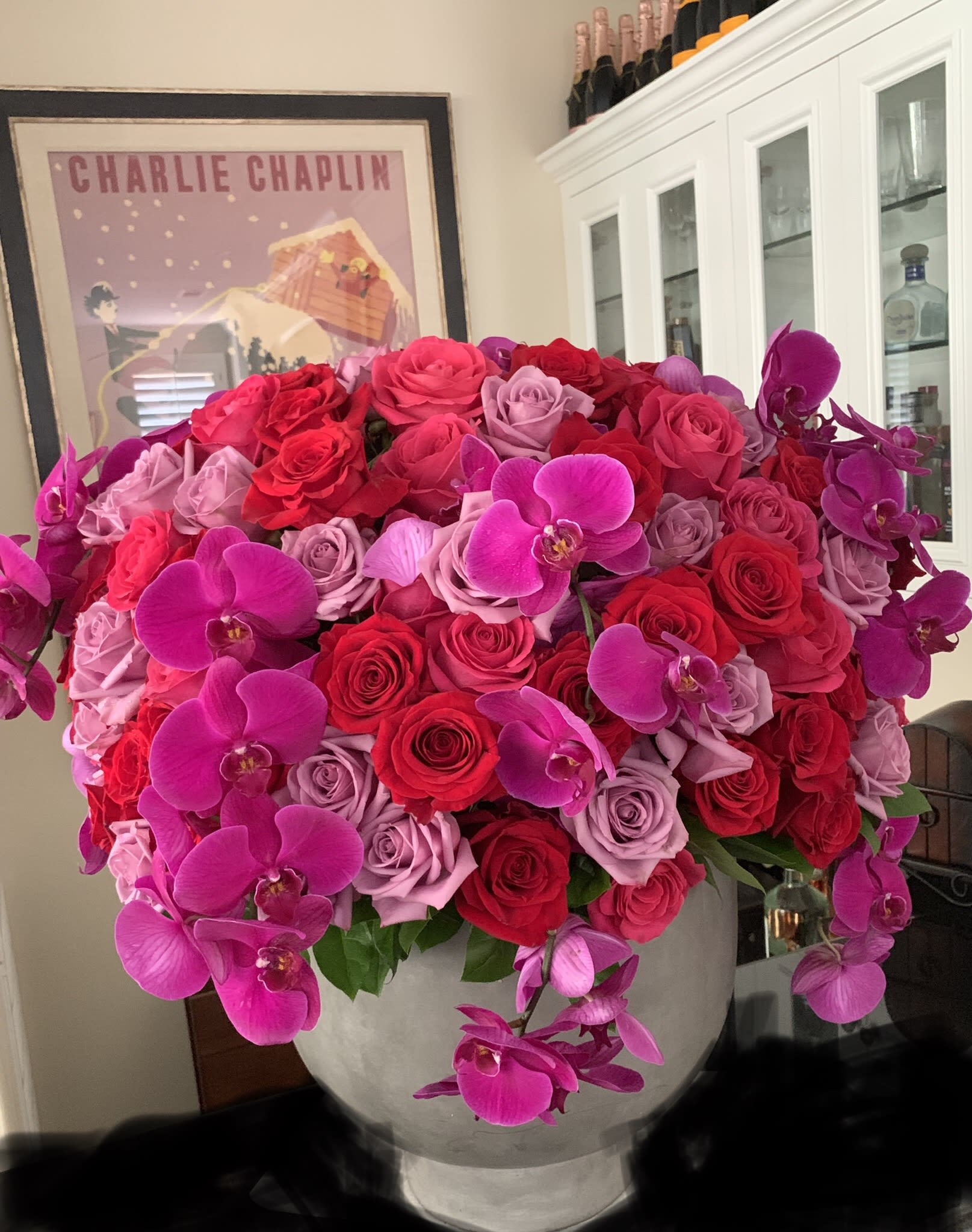 Large arrangement of red and pink roses with magenta orchids in a round white vase.