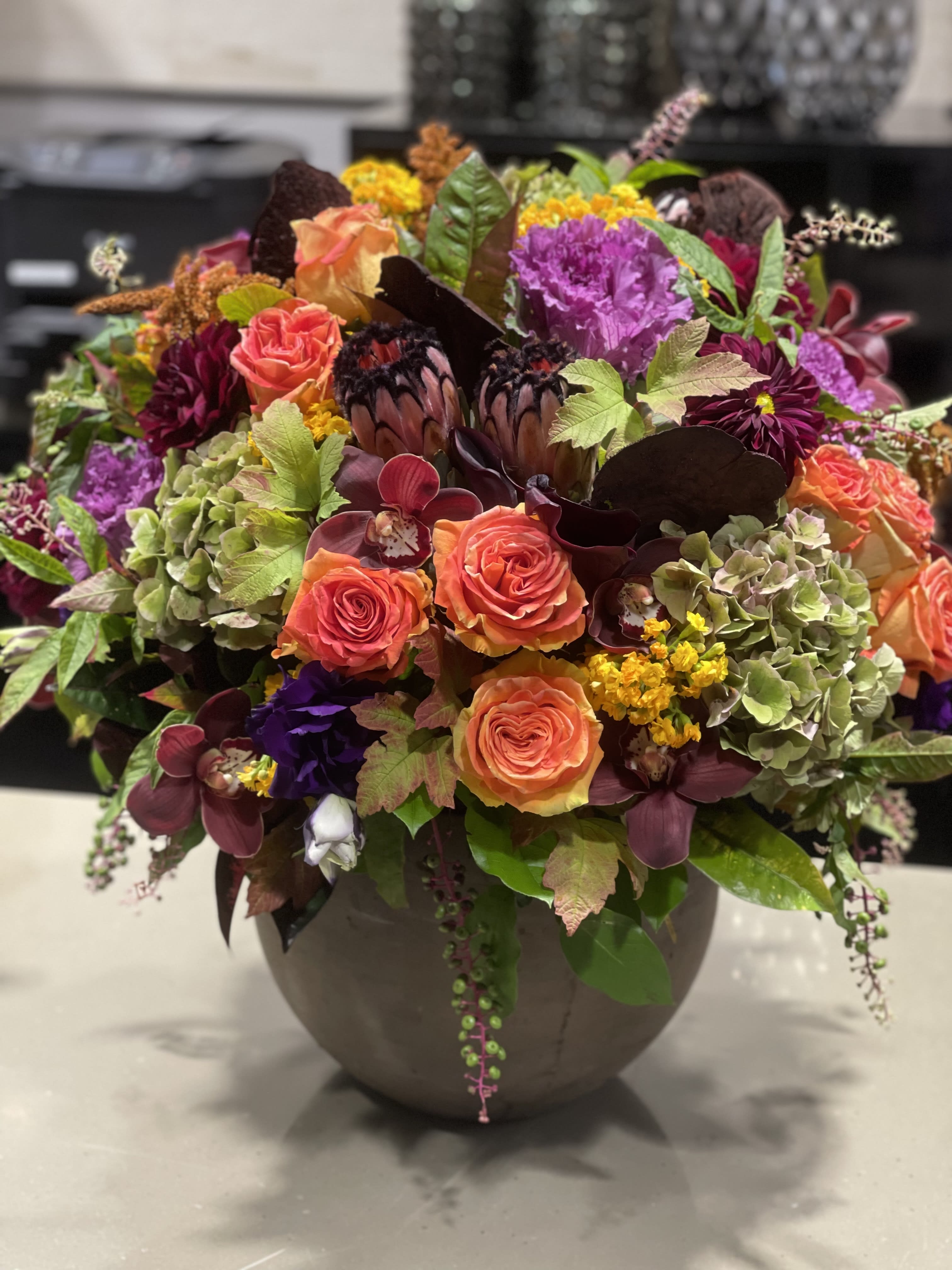 Lush autumn-toned arrangement with orange roses, protea, orchids and mixed blooms in a round ceramic vase