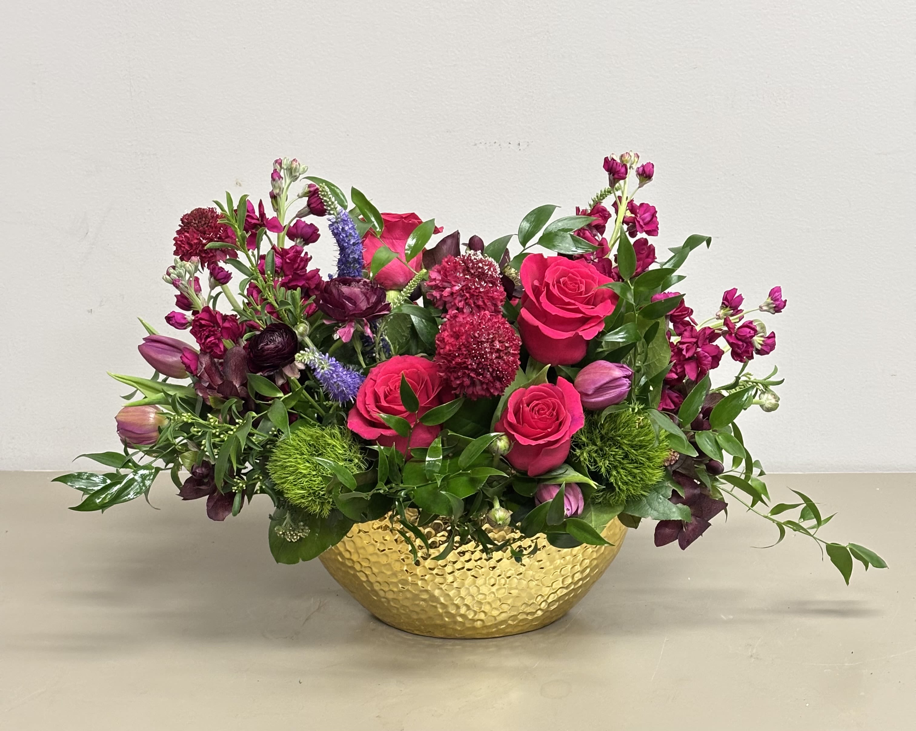 Low arrangement of hot pink roses and mixed purple flowers in a hammered gold bowl.