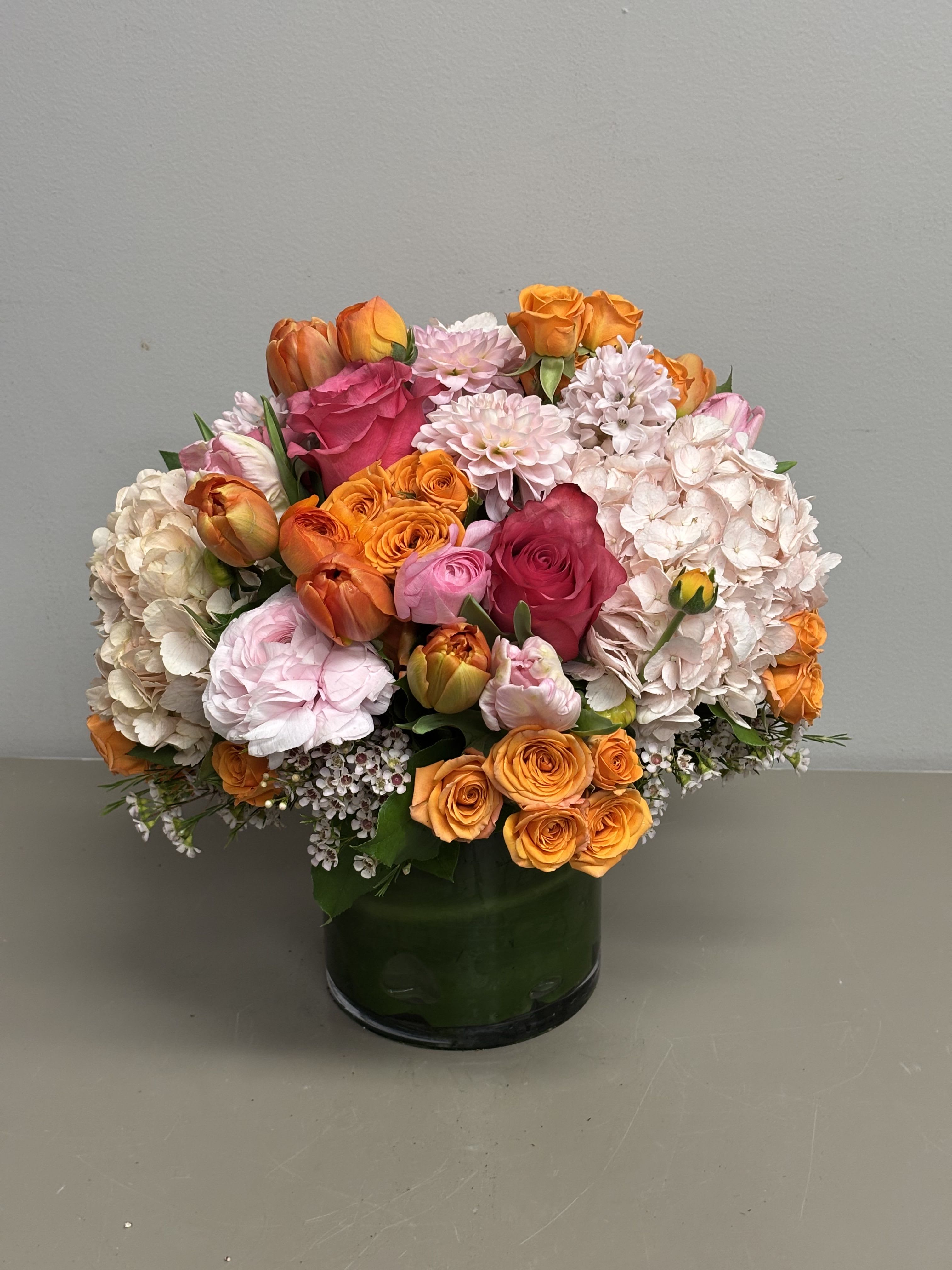 Radiance - A Beautiful combination of Bright colors of orange and pinks Hydrangeas, Roses, Spray Roses, Ranunculus, Tulips and other seasonal flowers in a glass cylinder lined with Ti leaves.