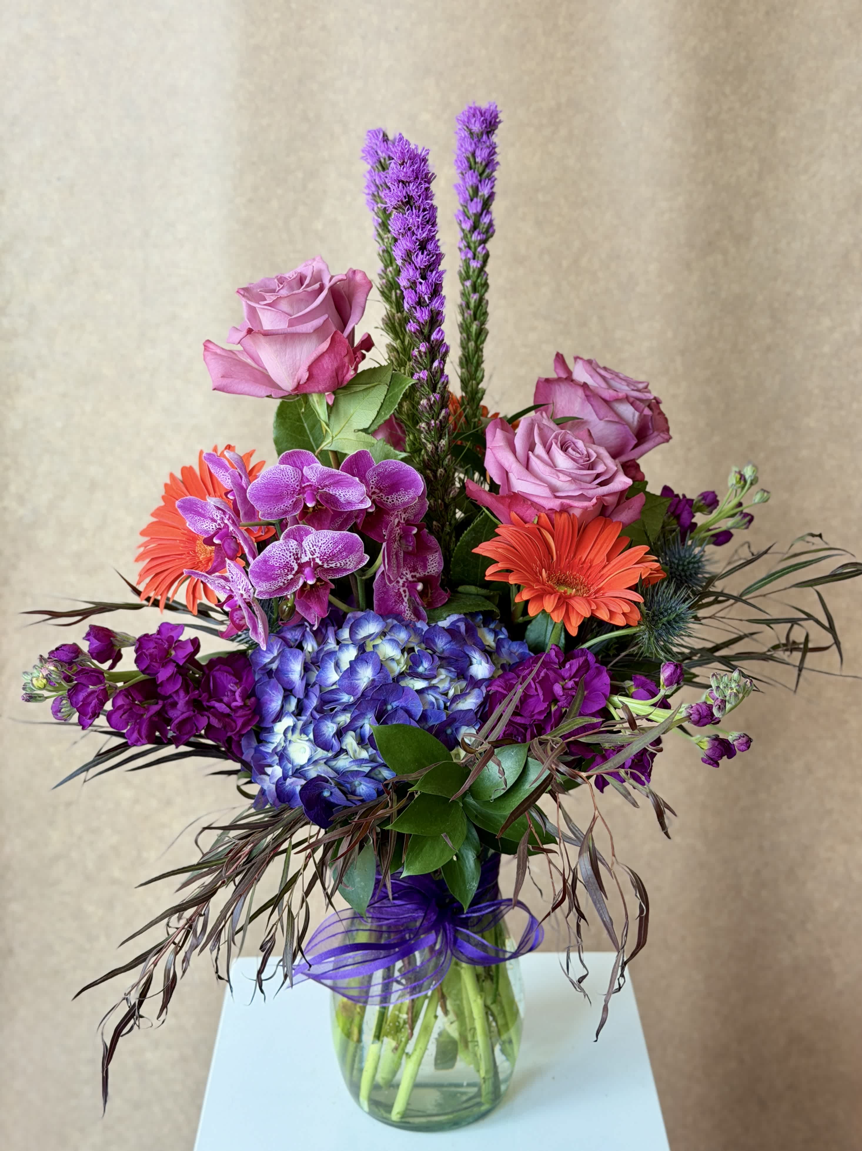 Mixed bouquet with pink roses, purple orchids, blue hydrangea, and orange gerbera daisies in a glass vase