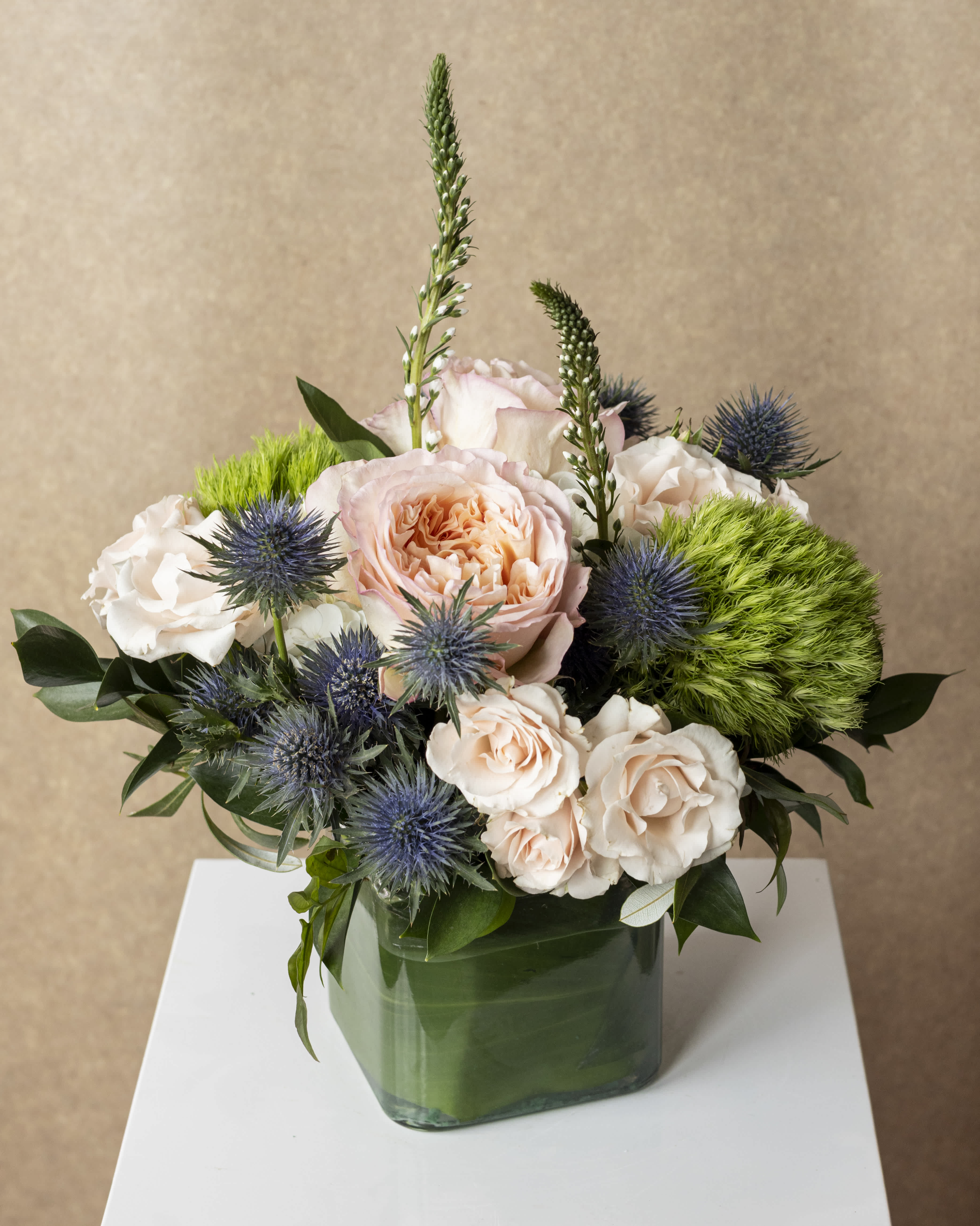 Peach and white roses in a green glass vase with blue thistle and green pom-pom flowers