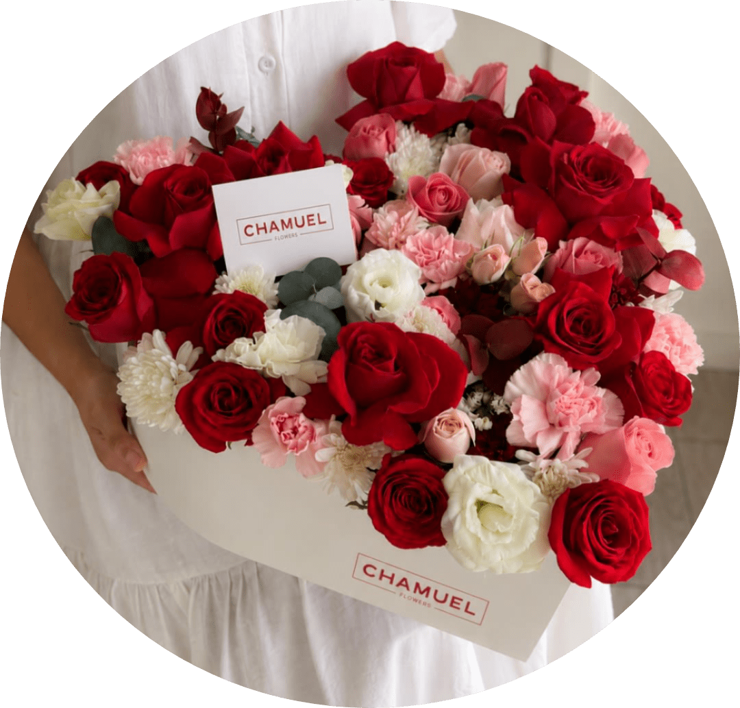 Heart-Shaped Box Mix Flowers Bouquet with Roses and Carnations - A lush heart-shaped arrangement of densely packed red roses opens like a declaration-perfect as an anniversary bouquet, a birthday surprise for someone you adore, or a "just because" gift to make them feel deeply seen. The HEART BOX MIX reads as love and romance at first glance: saturated red rose heads mingle with soft pink roses and carnations, while white blooms and tiny filler flowers catch the eye and soften the silhouette for an elegant, celebratory feel.  This boxed presentation comes in a cream-colored heart-shaped cardboard flower box with a visible branded card, giving the arrangement a polished, gift-ready look that arrives ready to impress. The mix includes roses, carnations, chrysanthemums, lisianthus, and small white accents, with muted foliage tucked throughout to add texture and depth. The dense, layered composition creates a luxurious, tactile surface that's both bold and tender.  Give this when you want the recipient to feel cherished-whether marking years together, celebrating a birthday, or sending a spontaneous reminder of affection. Sizes and care follow standard boxed-arrangement practices: keep blooms in a cool spot away from direct sun and change the water if provided; the heart box and printed logo card make gifting effortless and memorable.