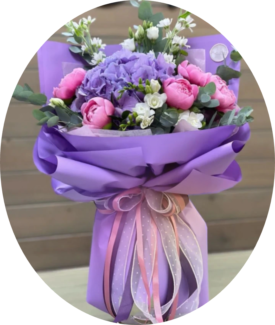 Lavender Hydrangea and Pink Peony Bouquet - A lavender hydrangea crowned with blushing pink peonies makes a soft, romantic statement in this hand-tied flower arrangement-perfect as a birthday bouquet, a tasteful anniversary gift, or a wedding-related presentation. The color story of purple, pink, white, and muted green reads springlike and feminine, the kind of floral gift that will make the person you're thinking of feel seen and cherished.  The bouquet features a large cluster of lavender hydrangea at its center, surrounded by peony-style pink blooms and delicate white freesia-like accents, with eucalyptus-like foliage adding gentle green contrast. It's wrapped in layered purple decorative paper and finished with long satin and sheer ribbon tails in pink and lavender for an elegant, polished look. The mix of textures-the fluffy hydrangea, lush peonies, and fine accent blooms-creates visual depth and a soft fragrance that complements the arrangement.  Ideal for celebrating milestones, saying "congratulations," or gifting on a romantic occasion, this arrangement arrives ready to present and makes a memorable centerpiece or bridal accessory. For best enjoyment, trim stems and change the water daily; available in a hand-tied size suited for gifting as shown.