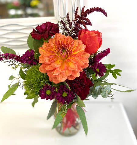 Compact bouquet with orange dahlia and rose, burgundy blooms, and green accents in a clear glass vase.