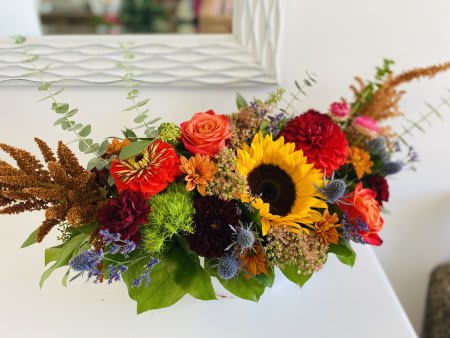 Marmalade Skies - Bring the warmth of the harvest season indoors with our Marmalade Skies centerpiece — a striking arrangement bursting with texture, color, and rustic charm. This elongated floral design features golden sunflowers, fiery orange roses, deep burgundy dahlias, velvety marigolds, and touches of burnt sienna amaranthus, all surrounded by wisps of eucalyptus and seasonal greenery.  Perfect for dining tables, mantels, or fall celebrations, Autumn Ember is a show-stopping piece that adds cozy elegance and a pop of seasonal brilliance to any space.