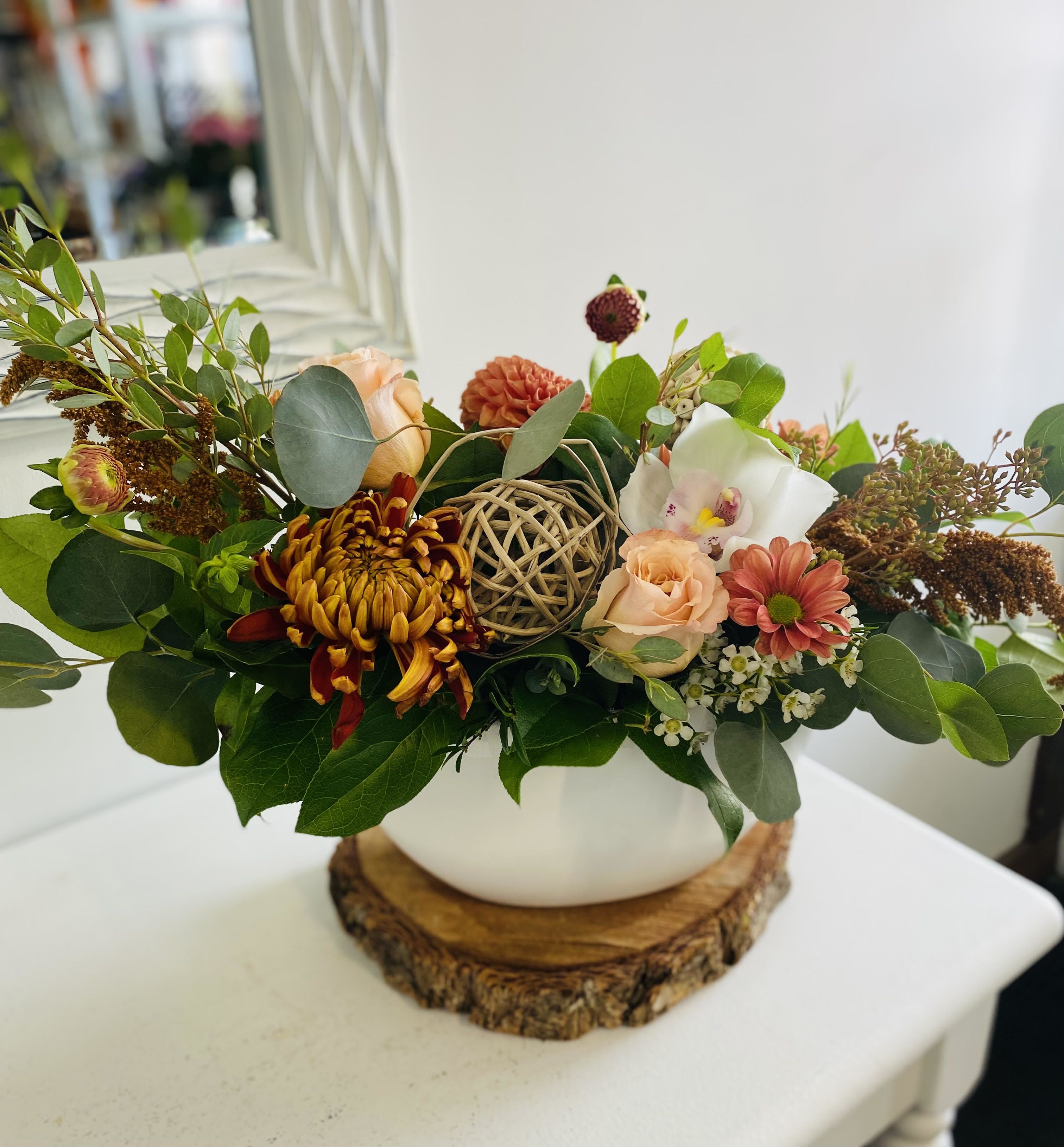 Low peach and rust flower arrangement with mums and orchid in a white bowl vase on a wood base