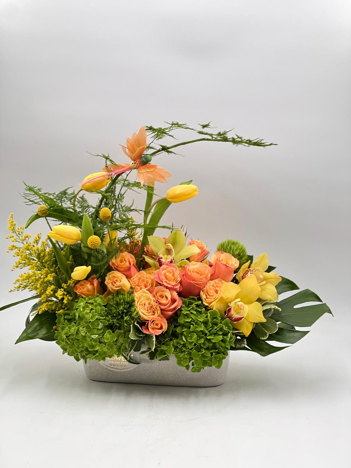 Pirouette - This vibrant, garden-style arrangement blends warm peach roses, sunny yellow tulips, and golden cymbidium orchids with lush green hydrangea and airy seasonal foliage. Designed with graceful height and movement, the composition feels light yet expressive, with stems gently arcing and flowers unfolding at different levels. Set in a low, modern rectangular vase, it brings a fresh, artistic energy that feels both cheerful and refined.  Perfect for brightening a room or marking a joyful occasion.