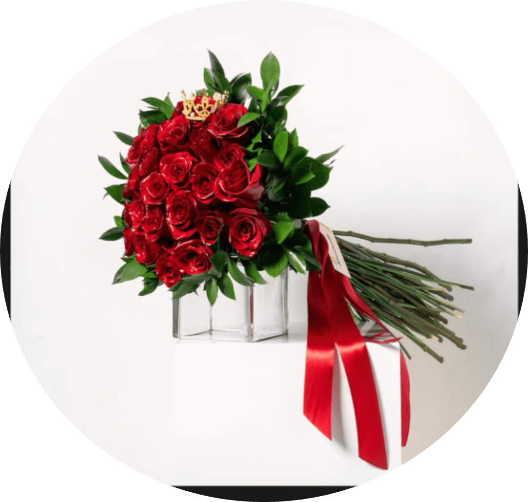 Nando Red Roses Bouquet in Clear Square Vase - A dense cluster of velvety red roses creates an instantly romantic centerpiece - perfect as an anniversary roses bouquet or a love-themed gift that says "you are cherished." The rounded spray of blooms presents a dramatic, formal silhouette that will make the person you're thinking of feel deeply celebrated and adored.  Glossy green foliage frames the saturated red petals for vivid contrast, arranged tightly in a clear square glass vase for a clean, modern finish. A red satin ribbon drapes from the side and a small gold crown pick adds a celebratory, luxurious accent, giving the arrangement a refined, regal touch.  Send this when you want to mark a milestone, rekindle romance, or make someone feel truly special; it reads as both intimate and grand. Vase-ready and arranged for lasting impact, place in fresh water away from direct heat and sunlight to help the roses maintain their look.