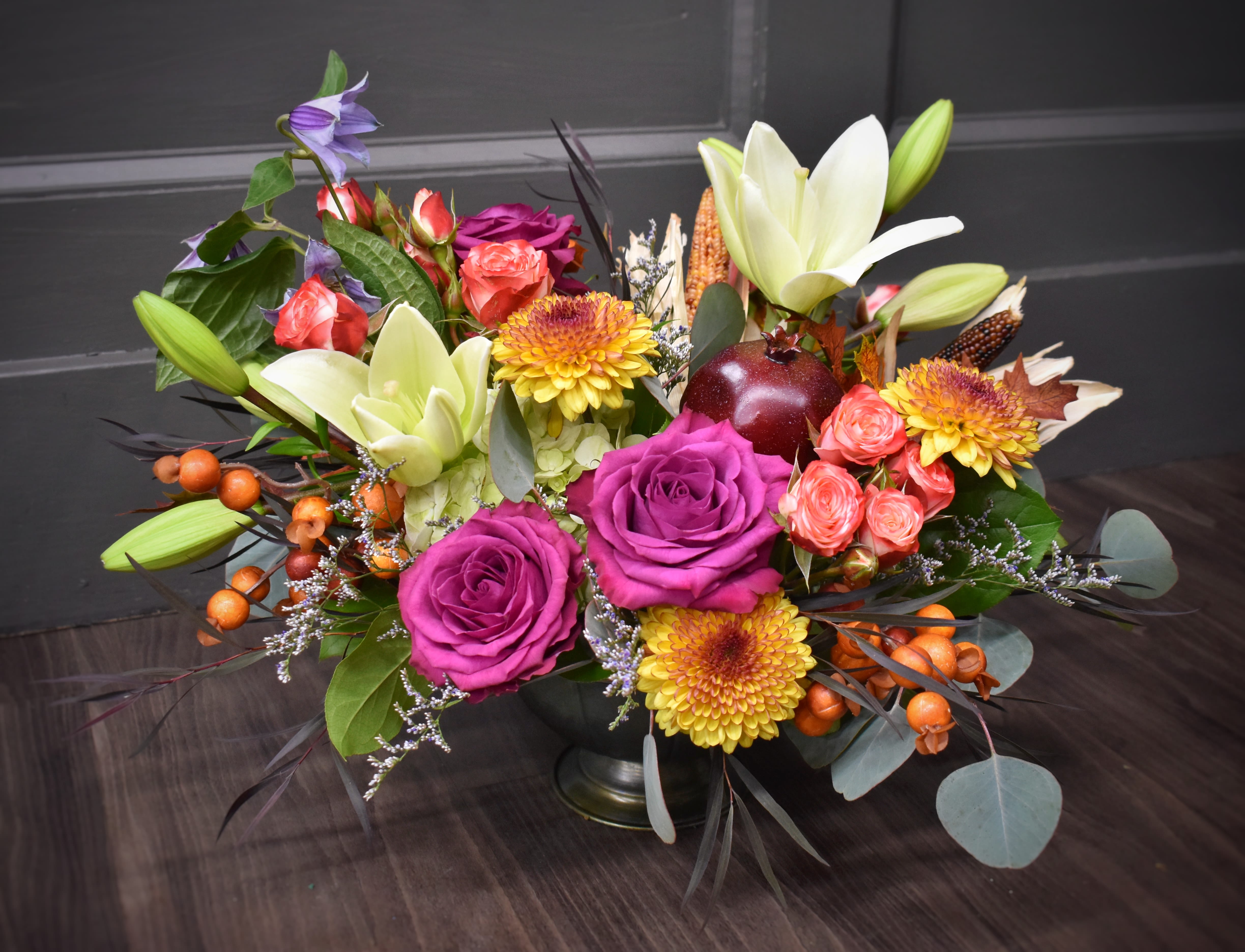 Premium Enchanted Harvest - Everything that reminds us of the harvest season, all in one luxurious compote. This arrangement is overflowing with a mix of hydrangea, roses, fall mums, dried corn, everlasting berries and a faux pomegranate. Our premium version is the most enchanted of them all and includes lilies and spray roses for a striking fall display. 