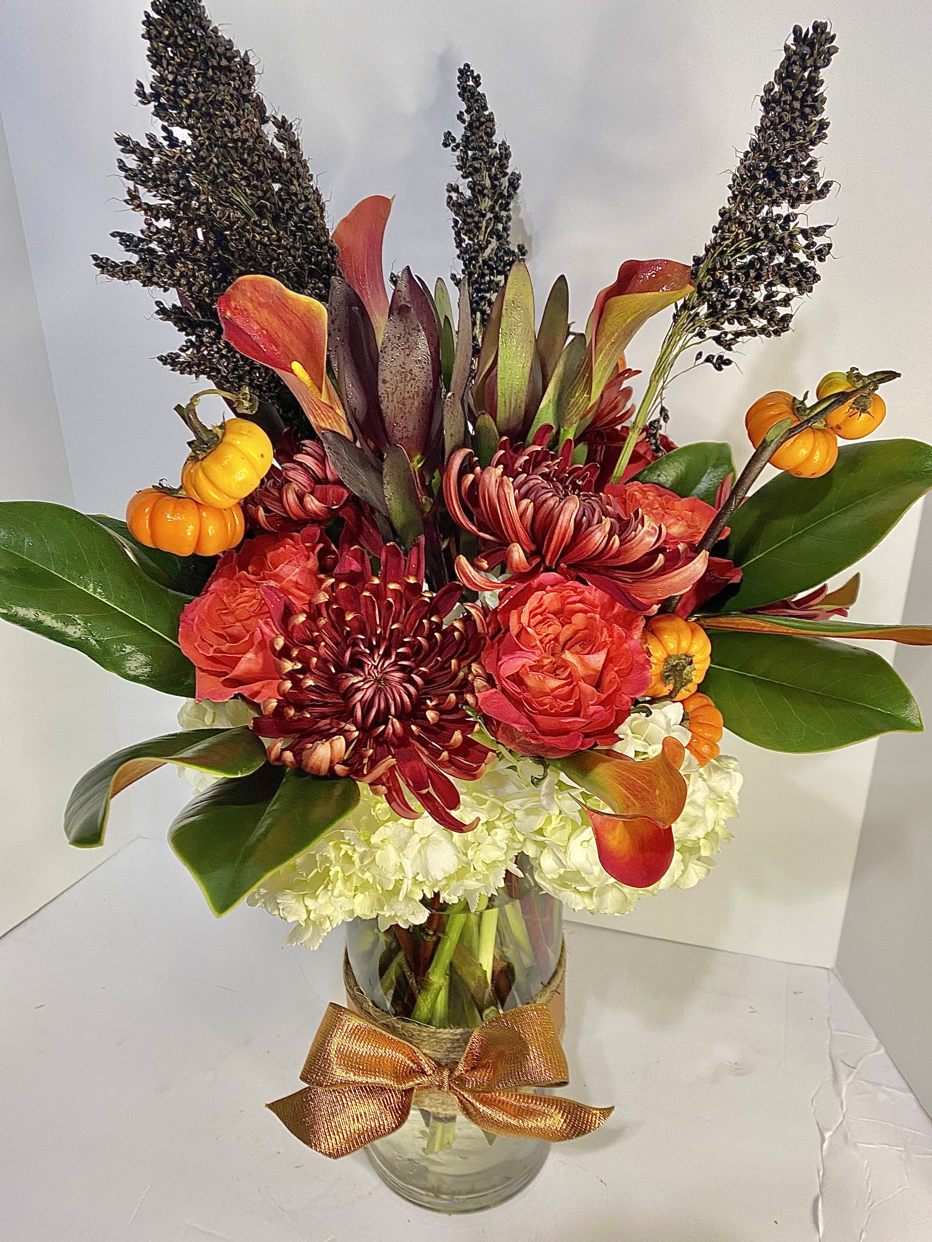 Tall fall arrangement with orange calla lilies, red mums, roses, and white hydrangeas in a glass vase with mini pumpkins