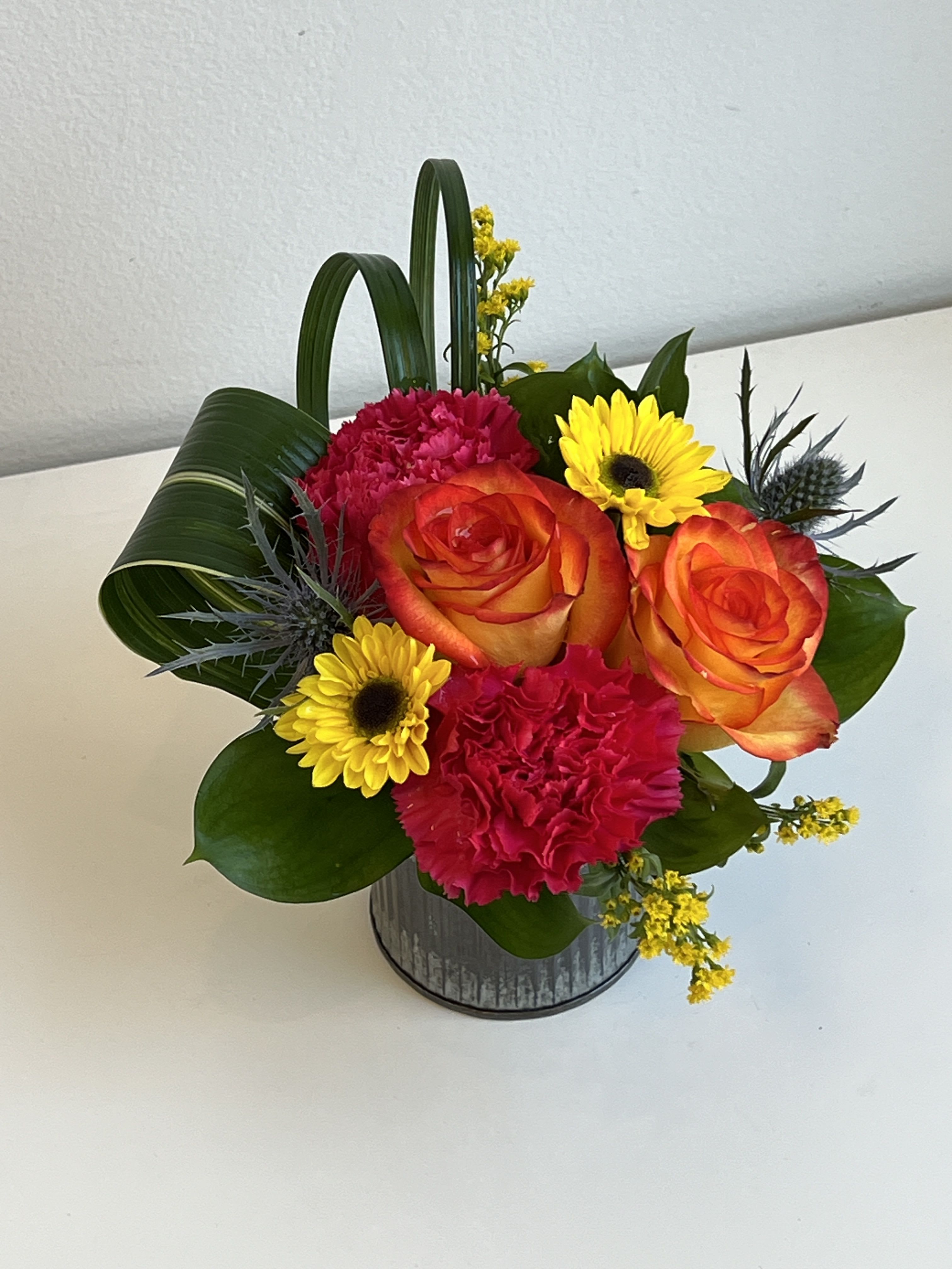 Bright bouquet of orange roses, pink carnations, and yellow daisies in a metal container