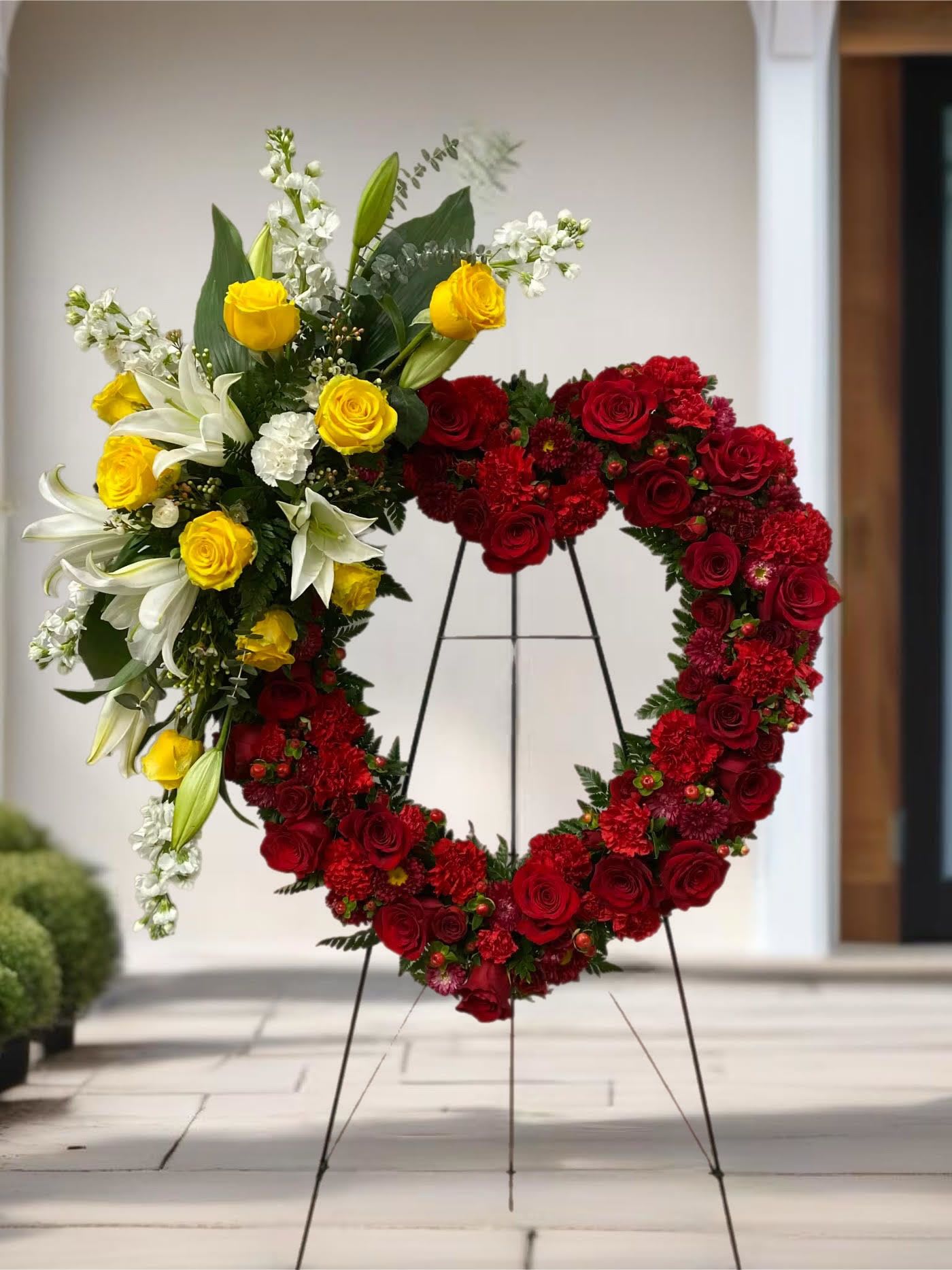 Heart-shaped floral wreath on a stand with red and yellow roses and white lilies