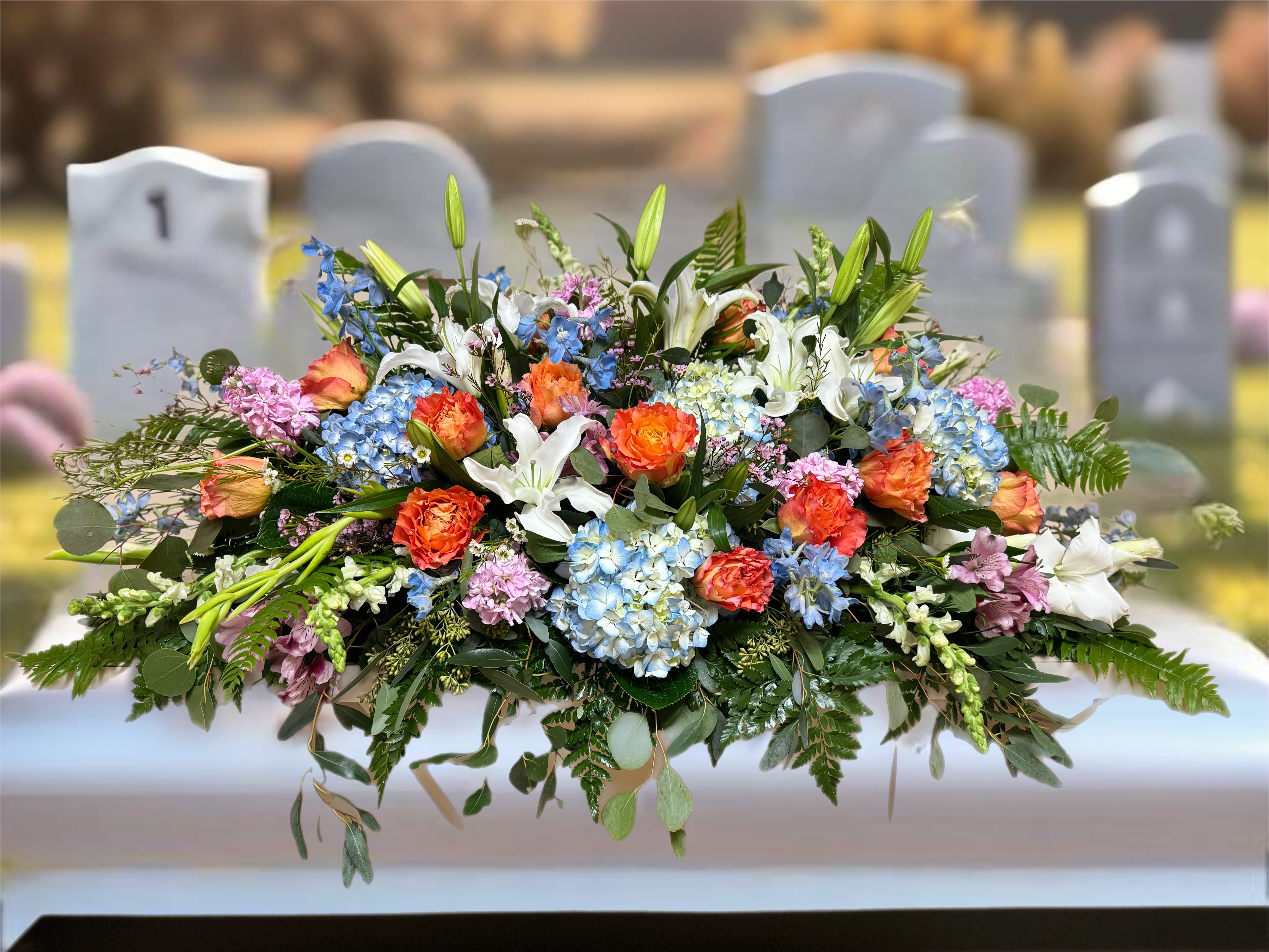Large mixed floral casket spray with lilies, roses, and blue hydrangeas