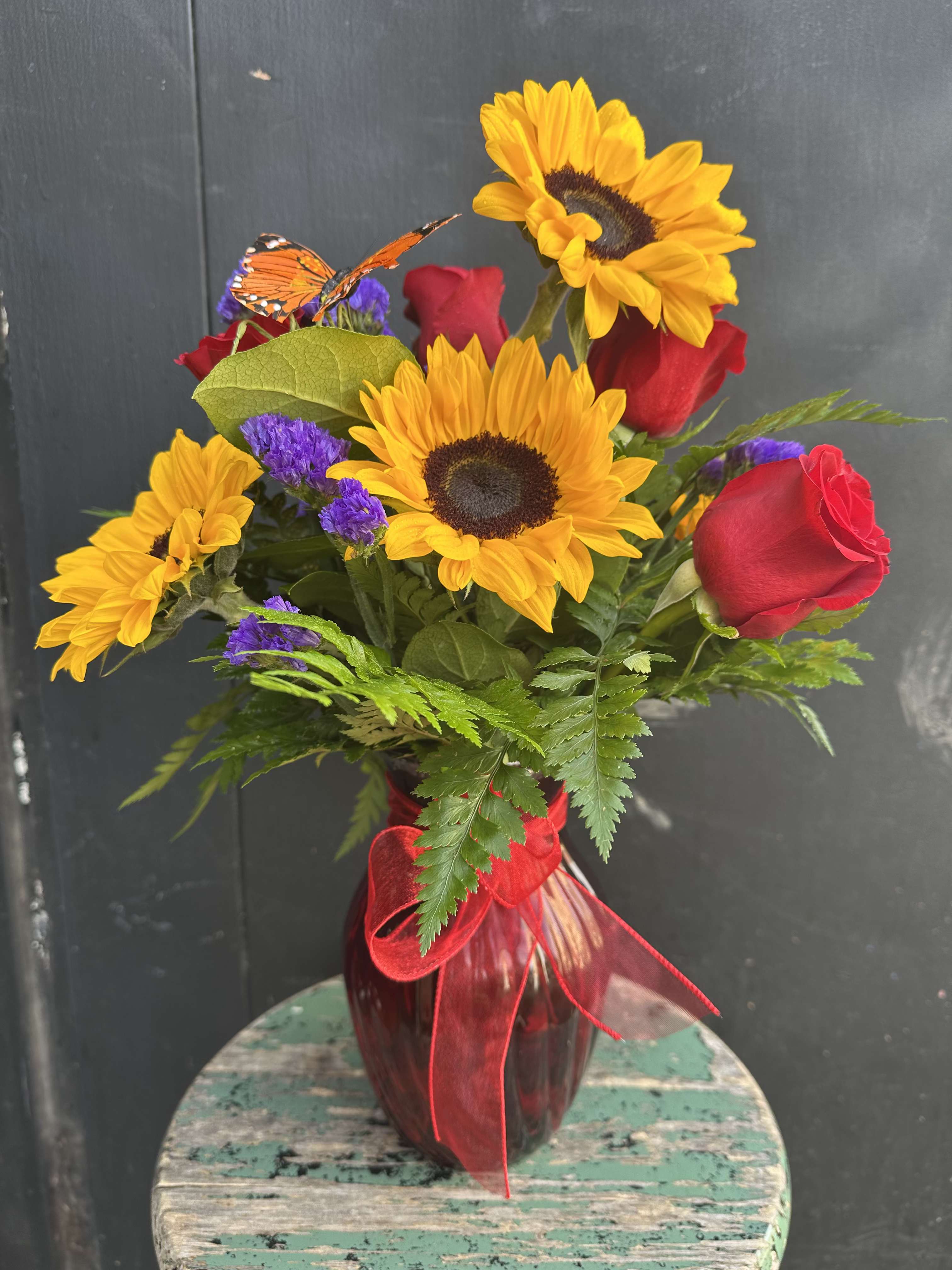 Butterfly Kisses Bouquet - Butterfly Kisses Bouquet is perfect for the Valentine who doesn't want a dozen red roses and enjoy wildflowers! This bouquet features: red roses, yellow sunflowers and purple statice and is in a ruby red ginger jar vase. 