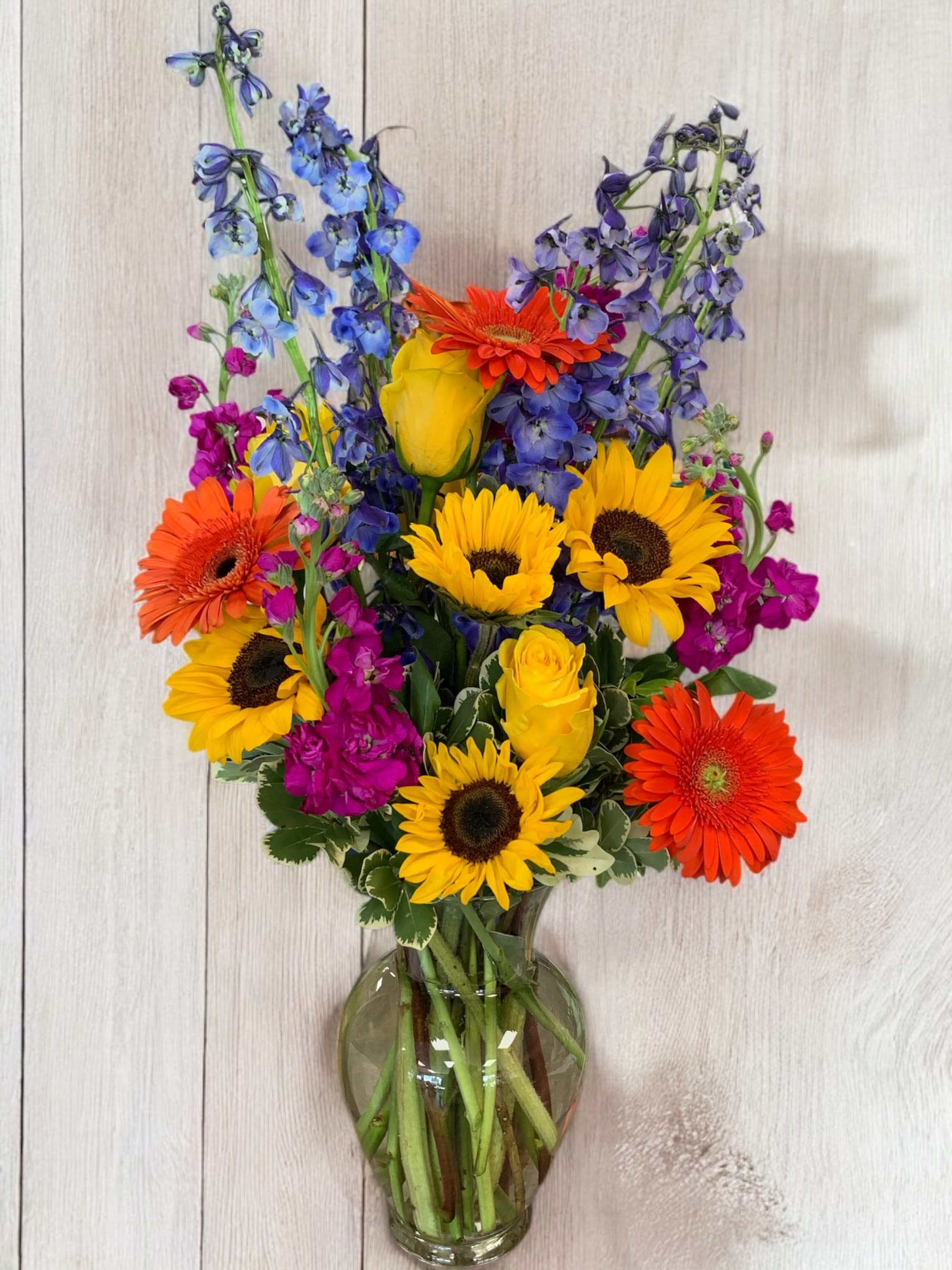 Bright mixed bouquet with sunflowers, gerberas, roses, and blue delphinium in a glass vase