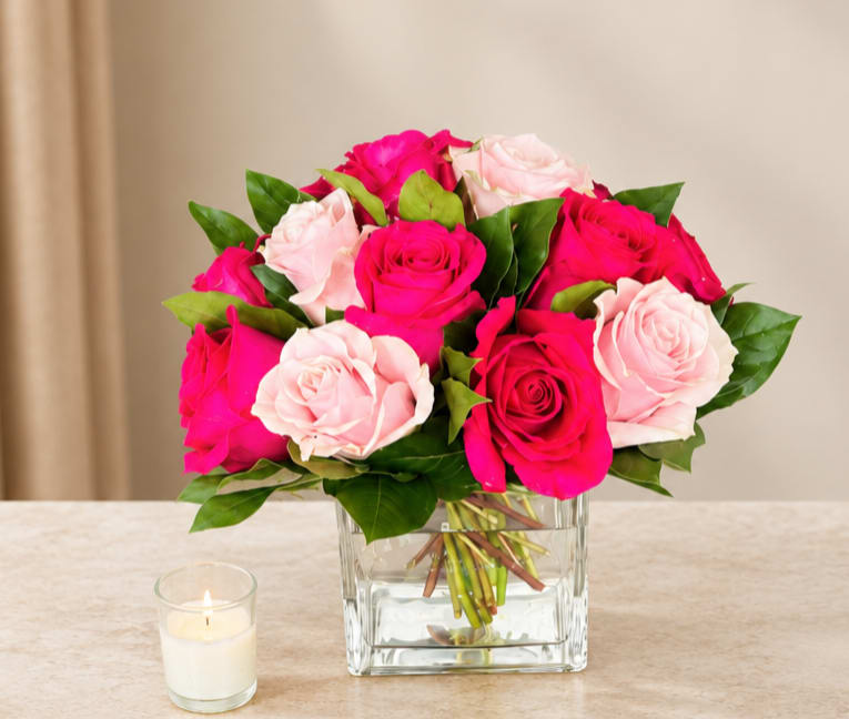 Pink and pale blush roses in a clear square vase beside a small candle