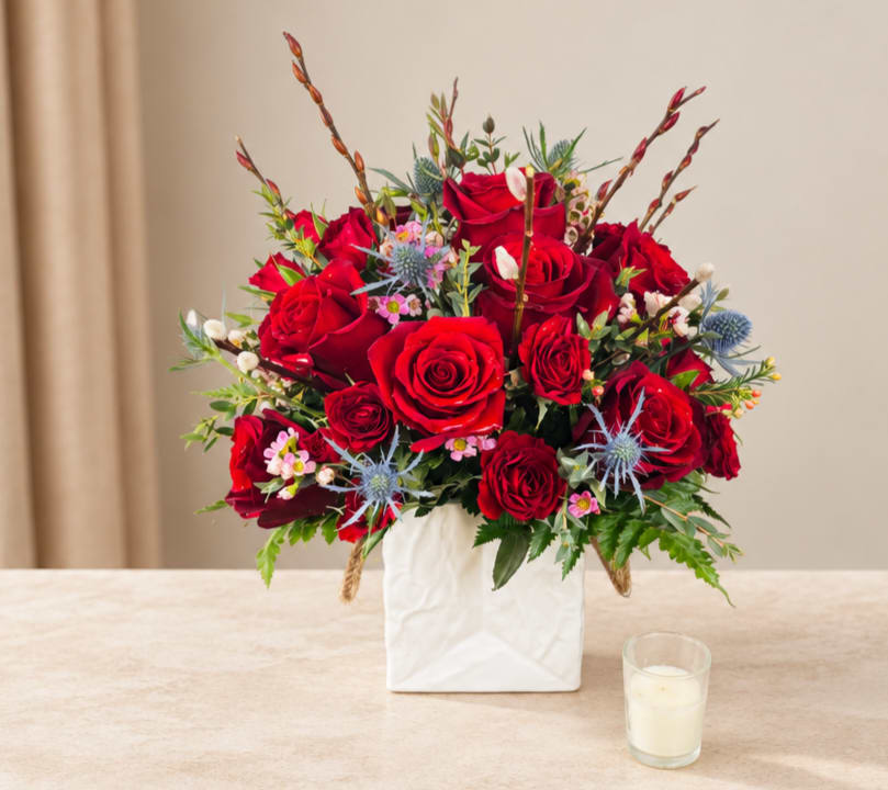 Red rose arrangement in a white square vase with small blue flowers