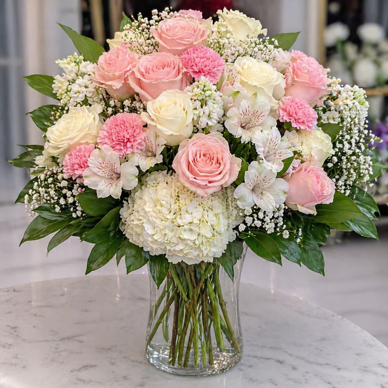 Rosalie Amour - Soft, romantic, and overflowing with graceful charm, Rosalie Amour is a lush luxury arrangement featuring blush pink roses, ivory hydrangeas, delicate alstroemeria, pink carnations, stock flower, and clouds of baby’s breath gathered in a classic clear glass vase. Its airy garden-inspired composition blends timeless romance with upscale floral artistry, creating a design that feels elegant, feminine, and effortlessly luxurious.