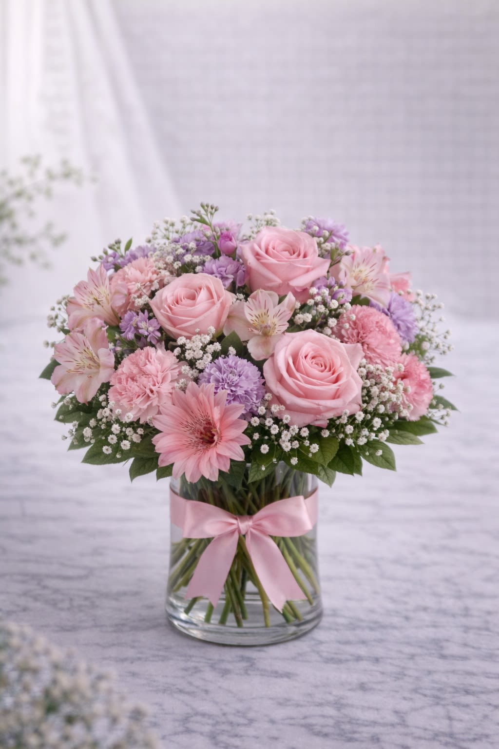 Rosewater Elegance - Blush Garden Whisper is a soft and romantic arrangement designed in delicate shades of blush pink and lavender. Featuring premium roses, carnations, alstroemeria, and seasonal accent blooms, this design feels airy, feminine, and effortlessly elegant. The clear glass vase is finished with a coordinating blush ribbon, creating a polished and graceful presentation. Perfect for birthdays, baby showers, anniversaries, or simply to brighten someone’s day, this arrangement delivers a gentle yet luxurious statement. Please note: Flower varieties may vary based on seasonal availability. If substitutions are necessary, we will always maintain the same blush pink and lavender color palette and overall design style. Containers and ribbon accents will always coordinate with the arrangement to ensure a cohesive and beautiful final look.