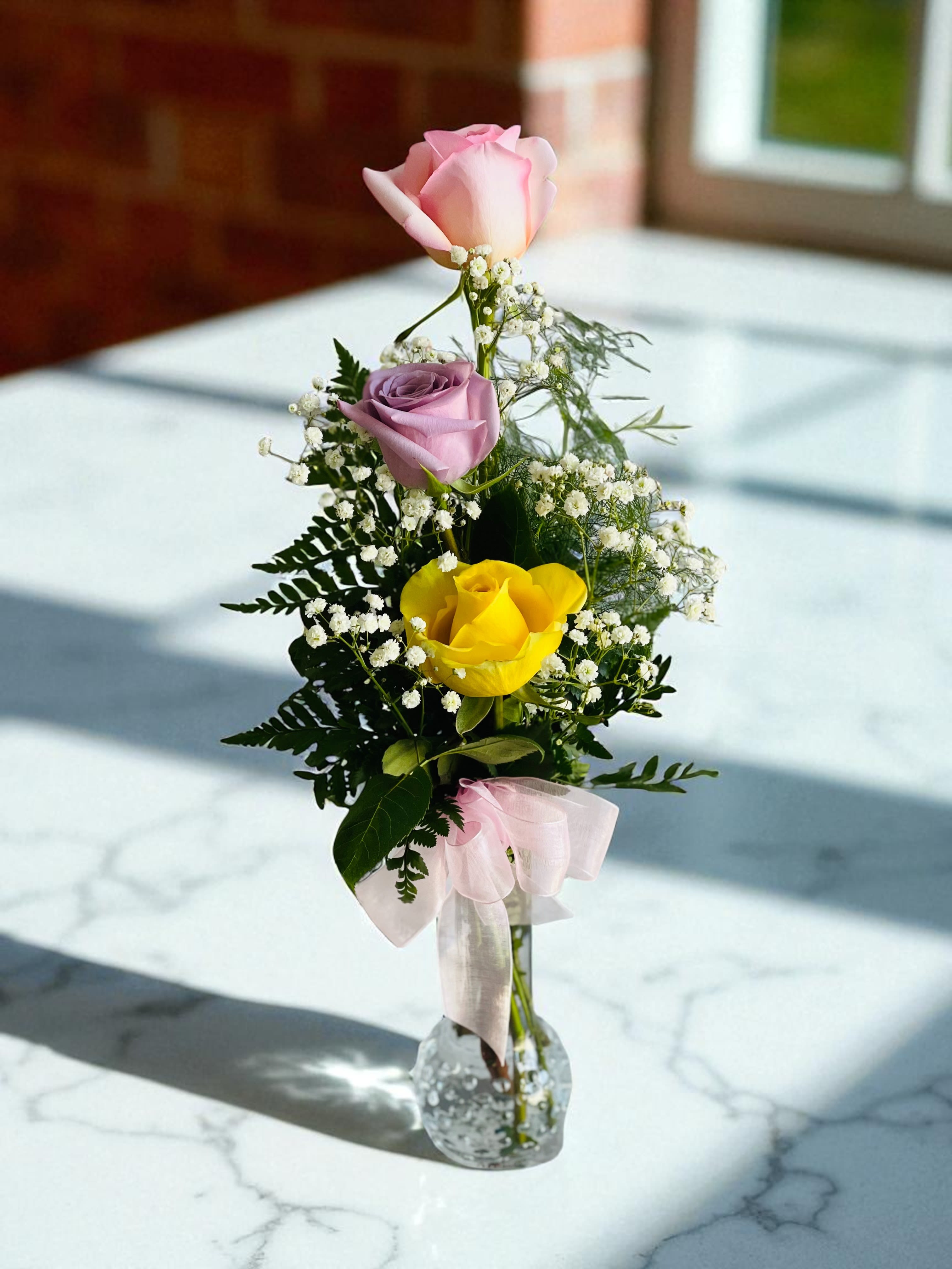 Pink, lavender, and yellow roses in a clear glass vase with a pink ribbon