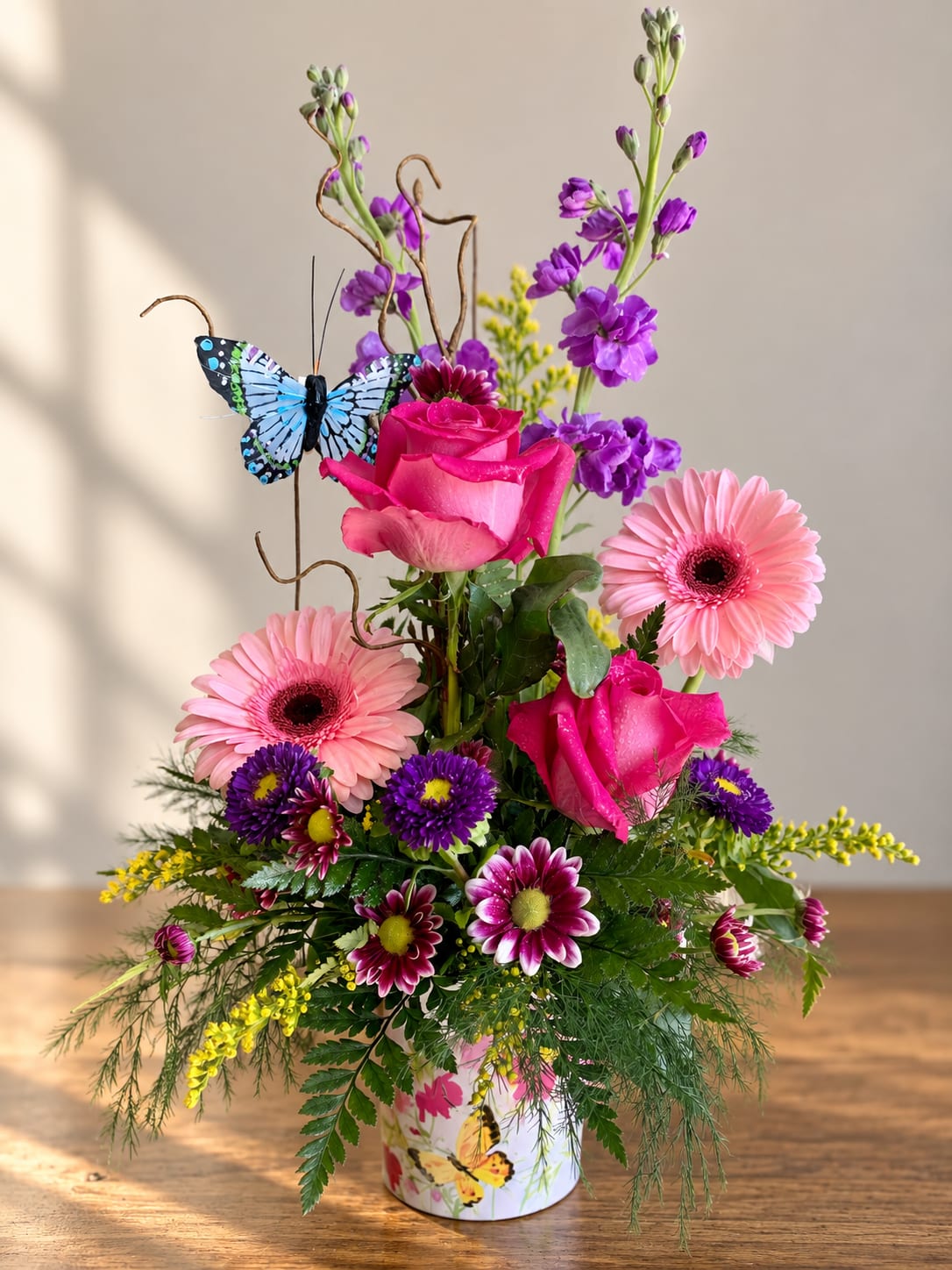 Pink roses and gerbera daisies in a butterfly-decorated vase with purple blooms