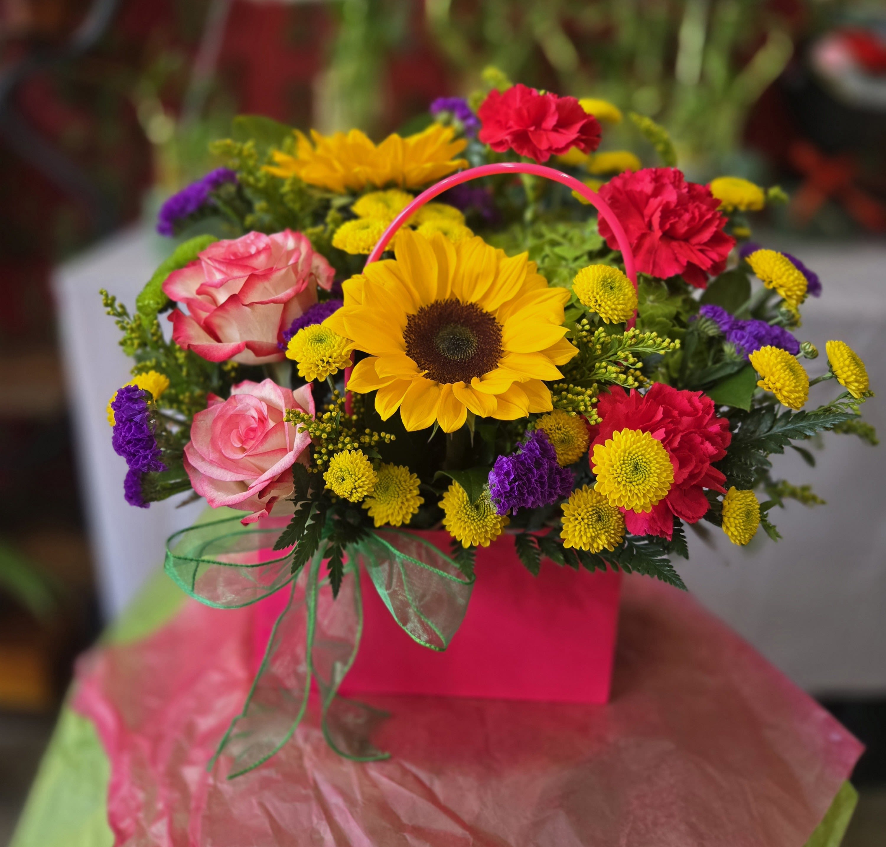 Mom's New Purse - A bright sunflower crowns the center of this cheerful flower arrangement-perfect as a birthday bouquet or "just because" gift to make Mom feel noticed. The arrangement reads like a celebratory purse-style gift box, so it works equally well for congratulations or a spontaneous pick-me-up for the person you're thinking of.  Pink roses and red carnations form a lush ring around the sunflower, with small yellow and purple chrysanthemums woven through for playful pops of color. The stems are arranged inside a pink handled gift box with a clear ribbon handle, and a sheer green ribbon is tied at the front to add a soft, ribboned finish. The overall effect is vibrant and lively: warm yellows and reds balanced by pretty pinks and cool purple accents.  This is the kind of gift that makes the recipient feel genuinely seen-bright, happy, and thoughtfully presented. Sized to sit on a table or bedside, it arrives ready to display in its boxed handle; change the water daily if you transfer the blooms to a vase, and order by the date you need it to ensure peak freshness.