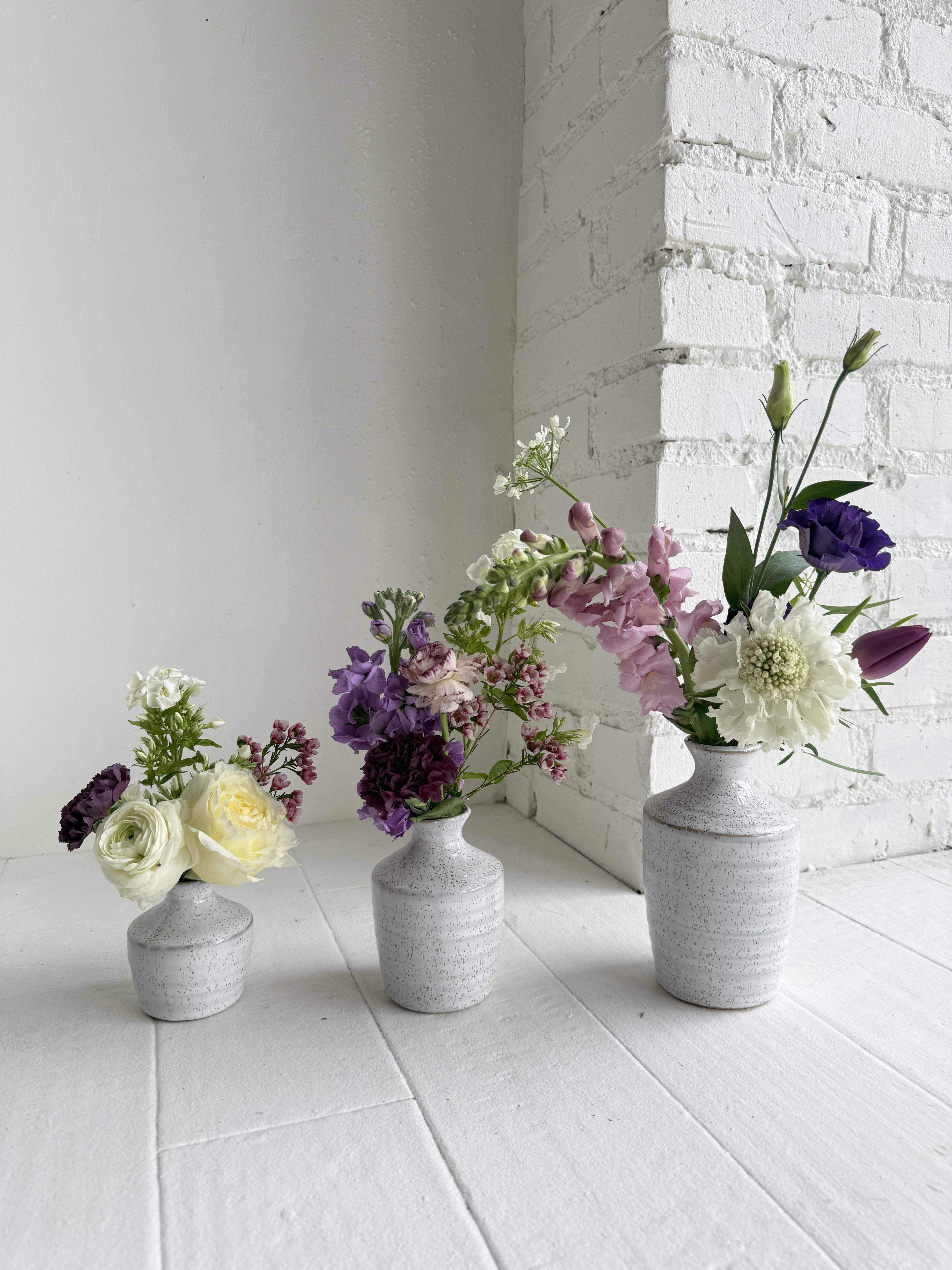Colette - Colette is a unique arrangement. We artfully arrange our favorite seasonal flowers in 3 vases that can be grouped together or divided and placed around the home or office for maximum enjoyment. All shades of purple are mixed with pops of creamy whites and greens.