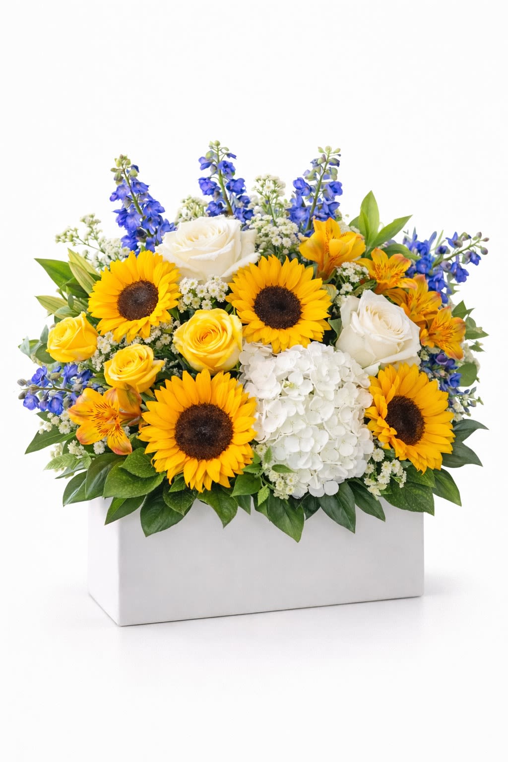 Sunny Meadow Mix - A vibrant and cheerful arrangement designed to brighten any day. Featuring radiant sunflowers, soft white and yellow roses, fresh white hydrangea, and seasonal accents in touches of blue and orange, this design brings a perfect balance of warmth and freshness. Arranged in a modern white container, it’s ideal for birthdays, get well wishes, thank you gifts, or simply to send a little sunshine. Flower varieties may vary slightly based on availability while maintaining the same color palette and overall style.