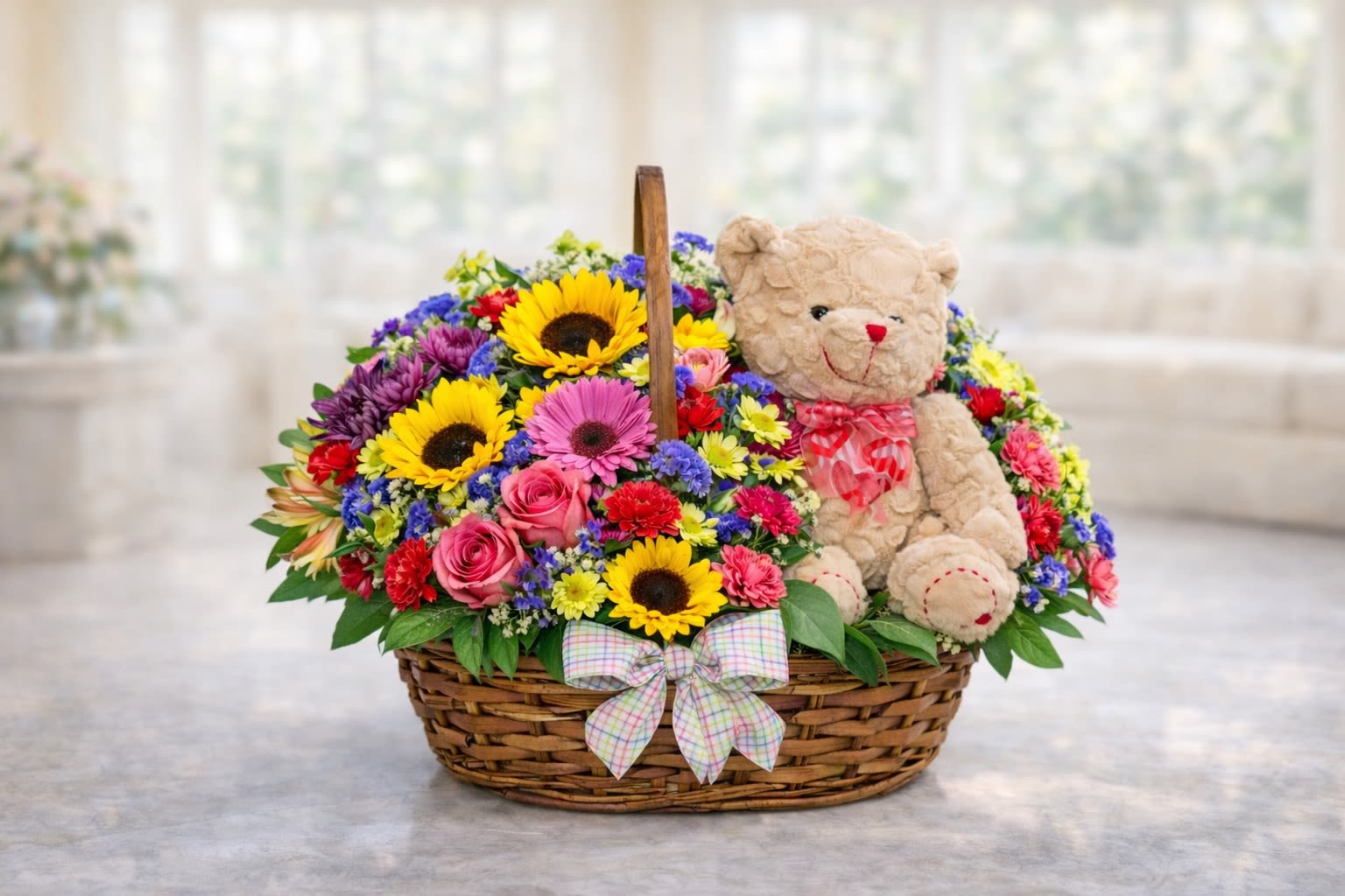 Sunny Smiles Gift Basket - A joyful and heartwarming basket arrangement featuring bright sunflowers, pink roses, colorful daisies, carnations, seasonal filler flowers, lush greenery, and an adorable plush teddy bear beautifully arranged in a rustic woven basket with decorative ribbon accents. This cheerful design is perfect for birthdays, get well wishes, celebrations, welcoming a new baby, or simply making someone smile.  Flower varieties, ribbon styles, and teddy bear design may vary slightly based on seasonal availability and inventory. In the rare event certain flowers or containers are unavailable, substitutions of equal or greater value will be used while maintaining the overall color palette and design style shown.