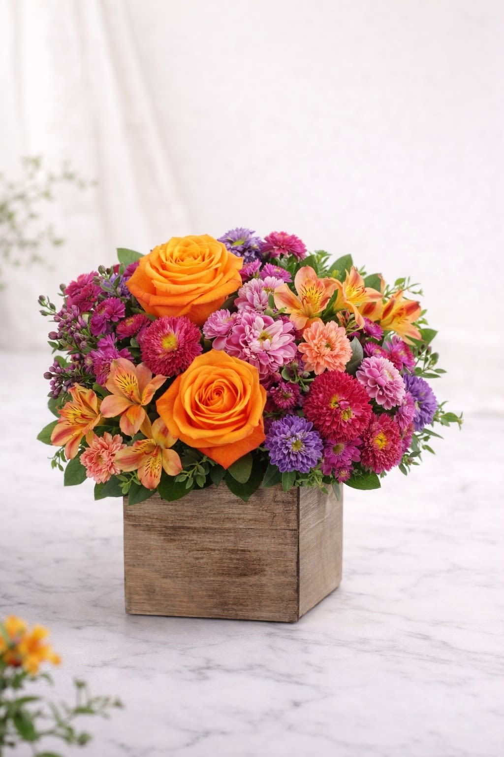 Sunset Celebration Cube - A bold and vibrant garden-style arrangement featuring bright orange roses, cheerful pink and purple blooms, soft accent flowers, and fresh greenery, beautifully designed in a rustic wooden cube container. This arrangement blends warm sunset tones of orange with playful pinks and purples, creating a joyful and energetic look that’s perfect for birthdays, congratulations, thank you gifts, or simply brightening someone’s day.  Substitution Policy: Flower varieties may vary depending on seasonal availability, but the overall color palette of orange, pink, and purple tones will always remain consistent. Any substitutions will be of equal or greater value and carefully selected to preserve the full, colorful garden style shown. ? Container Notice: The wooden cube container may vary slightly based on availability, but it will always complement the arrangement and match the warm, natural aesthetic. Our goal is to deliver an arrangement that reflects the same vibrancy, fullness, and joyful color harmony as pictured.