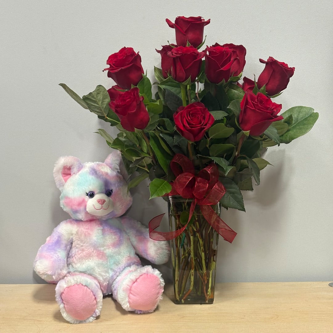 Red roses in a glass vase beside a pastel teddy bear