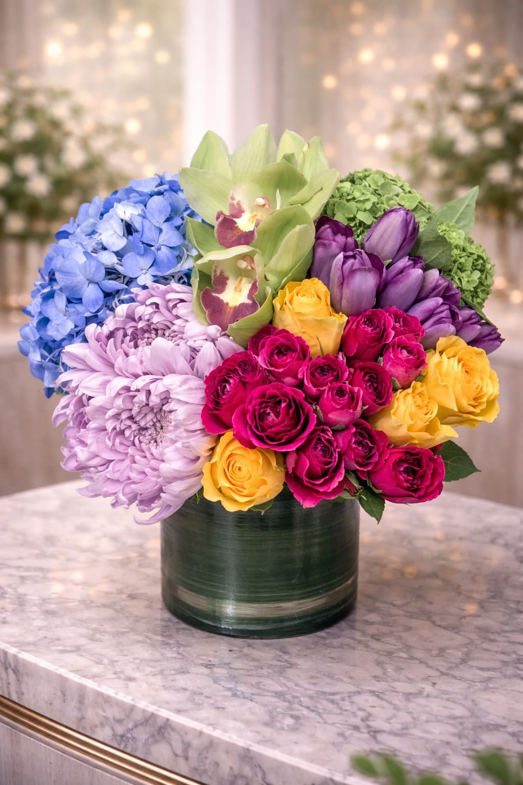 The Grand Color Symphony - A bold and joyful luxury arrangement featuring premium green cymbidium orchids, lush hydrangea, vibrant garden roses, elegant tulips, and full chrysanthemum blooms. Designed in a modern glass cylinder, this piece blends rich jewel tones — fuchsia, golden yellow, lavender, emerald green, and soft blue — creating a striking yet balanced garden-inspired centerpiece. Perfect for birthdays, congratulations, celebrations, thank-you gifts, or anyone who loves color and statement florals. • Premium green cymbidium orchids • Blue and green hydrangea • Garden roses in fuchsia &amp; yellow • Purple tulips • Full chrysanthemum bloom • Modern glass vase included This arrangement is full, textured, and designed to make an unforgettable impression. ⸻  Seasonal &amp; Freshness Guarantee We select the freshest seasonal blooms available to ensure vibrant color and long-lasting beauty. Because flowers are seasonal, certain varieties may occasionally be substituted. If substitutions are necessary, we will: • Maintain the same bright, colorful palette • Preserve the full, lush garden style • Keep the same premium value and size • Use flowers of equal or greater quality Your arrangement will always reflect the same vibrant, celebratory look and overall design style shown — never a completely different color theme or aesthetic. Each piece is handcrafted with care to ensure fullness, balance, and beauty.