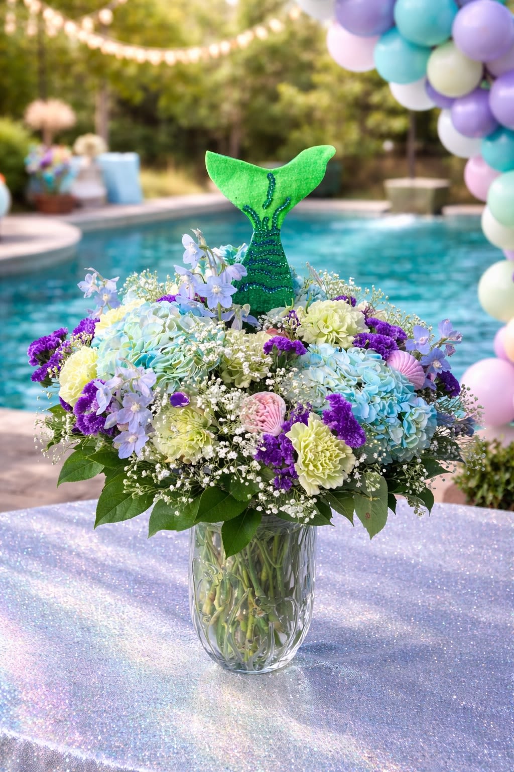 Under the Sea Celebration - A whimsical and enchanting arrangement inspired by an underwater garden, featuring lush blue hydrangeas, soft pastel blooms, delicate filler flowers, and a sparkling mermaid tail accent. Designed in a clear glass vase, this piece blends ocean-inspired tones of aqua, lavender, soft green, and white for a magical and playful presentation. The mermaid tail detail adds a fun and festive touch, making it perfect for themed celebrations. Perfect for birthdays, mermaid-themed parties, baby showers, poolside events, or anyone who loves a little magic. • Premium blue hydrangeas • Soft pastel seasonal blooms • Delicate filler accents • Decorative mermaid tail topper • Clear glass vase included Handcrafted for fullness, color harmony, and celebration impact. ⸻  Seasonal &amp; Freshness Guarantee We use the freshest seasonal blooms available to ensure beauty and longevity. Because flowers and decorative accents are seasonal, certain blooms or shades may occasionally be substituted. If substitutions are necessary, we will: • Maintain the same ocean-inspired color palette (blues, purples, soft greens) • Preserve the same full, festive design style • Keep the same overall size and decorative impact • Use flowers and accents of equal or greater quality Your arrangement will always reflect the same playful, magical aesthetic shown — never a different theme or simplified version. Each piece is handcrafted to ensure balance, fullness, and celebration-ready presentation.